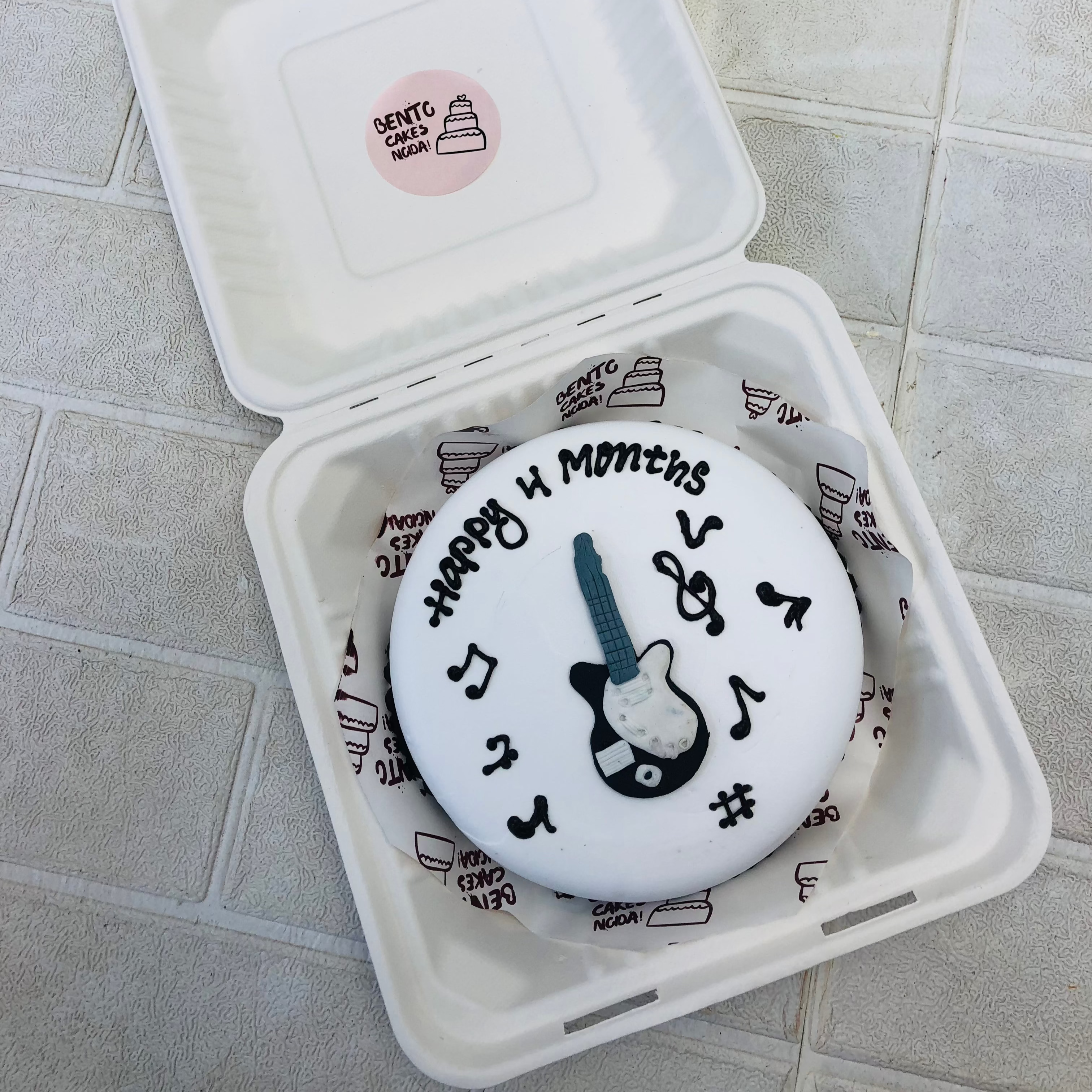 A white cake in bento box with a Guitar design and music symbols on it for 4 months baby.