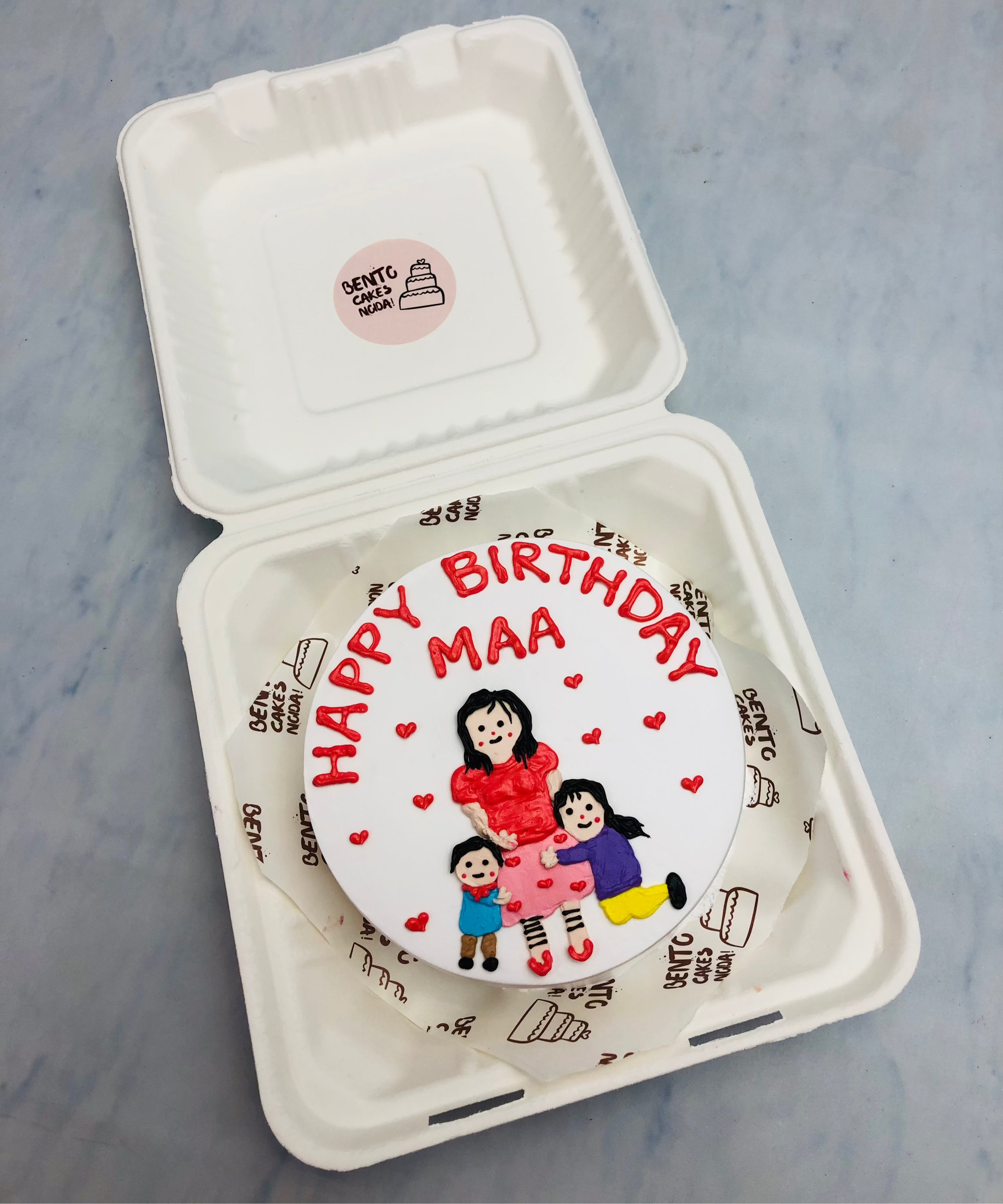 A heartfelt birthday cake for a mother, adorned with a fondant illustration of a mother and two children, surrounded by small red hearts. The cake features the text "HAPPY BIRTHDAY MAA" in bold red letters.