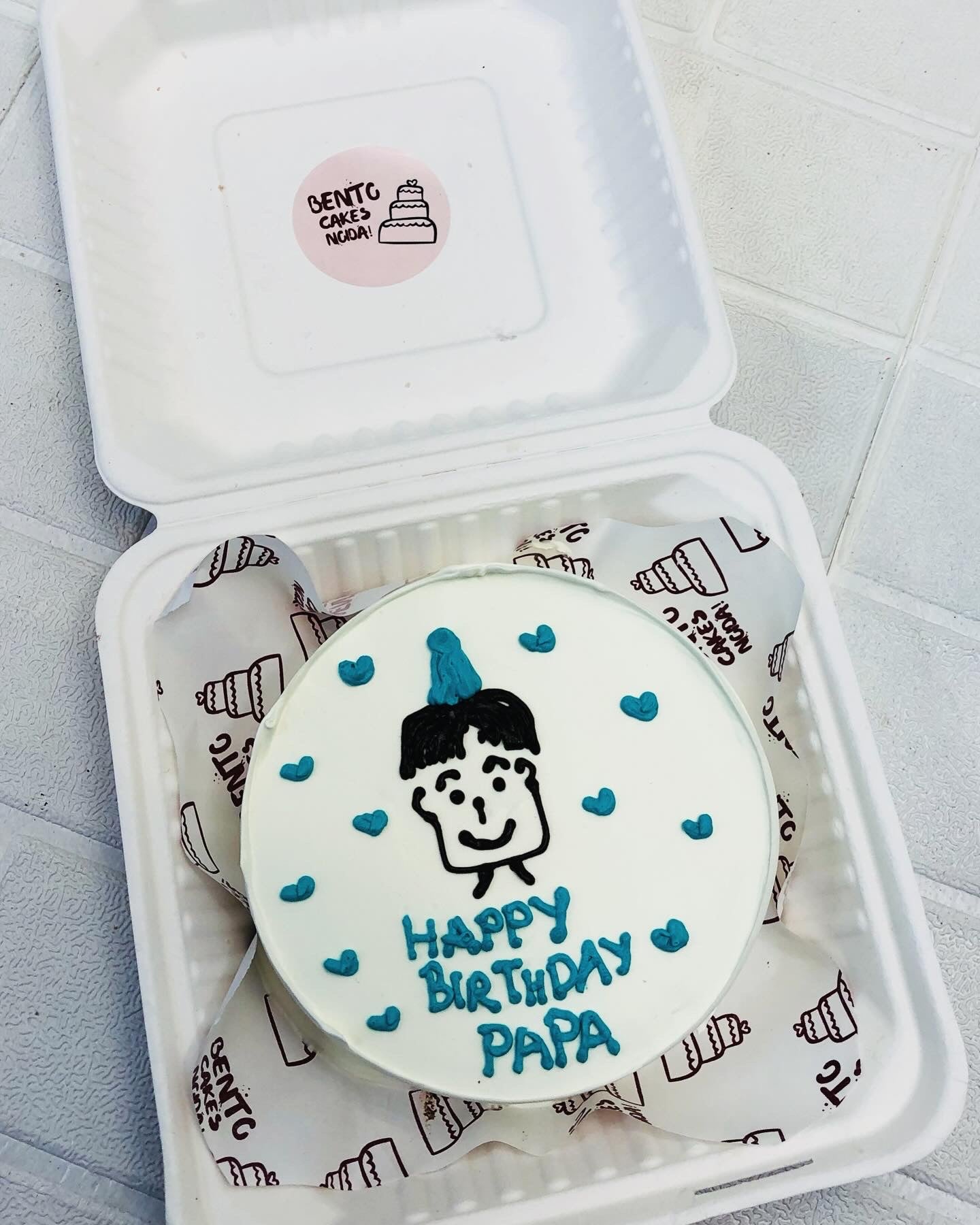 A white birthday cake with blue decorations, featuring a cartoon drawing of a person wearing a party hat. The cake has 'HAPPY BIRTHDAY PAPA' written in blue icing and is inside a bento box.