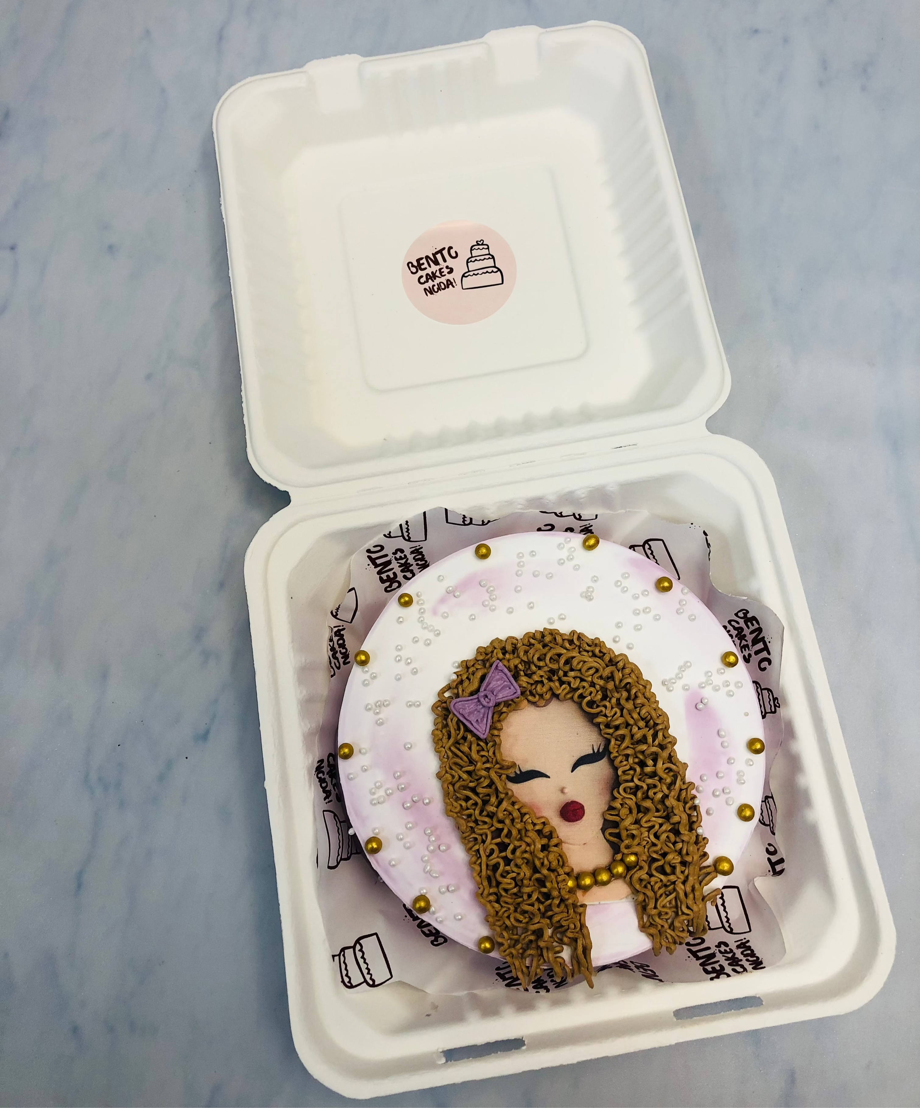 A beautifully decorated bento cake inside a bento box, featuring an artistic design of a woman's face with curly brown hair, a purple bow, and bold red lips. The cake is adorned with delicate white and gold pearls, adding a touch of elegance. 