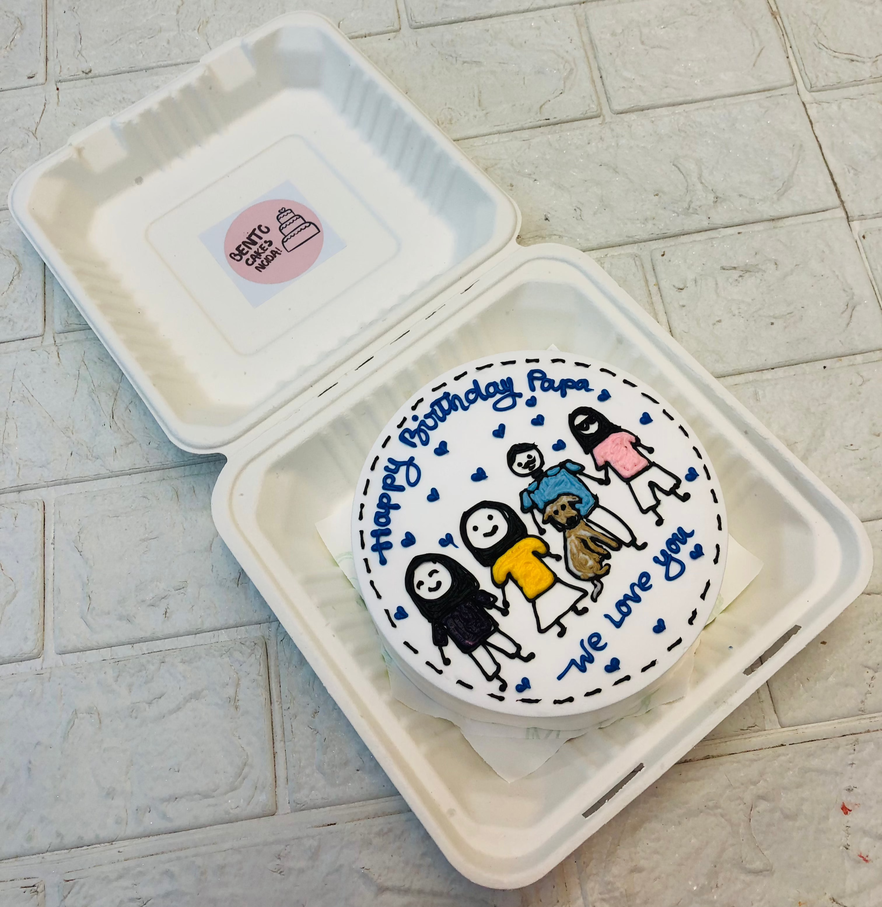 A custom birthday cake inside a takeout box, featuring a hand-drawn illustration of four family members and a dog. The cake is decorated with heartfelt blue icing messages: "Happy Birthday Papa" and "We love you."