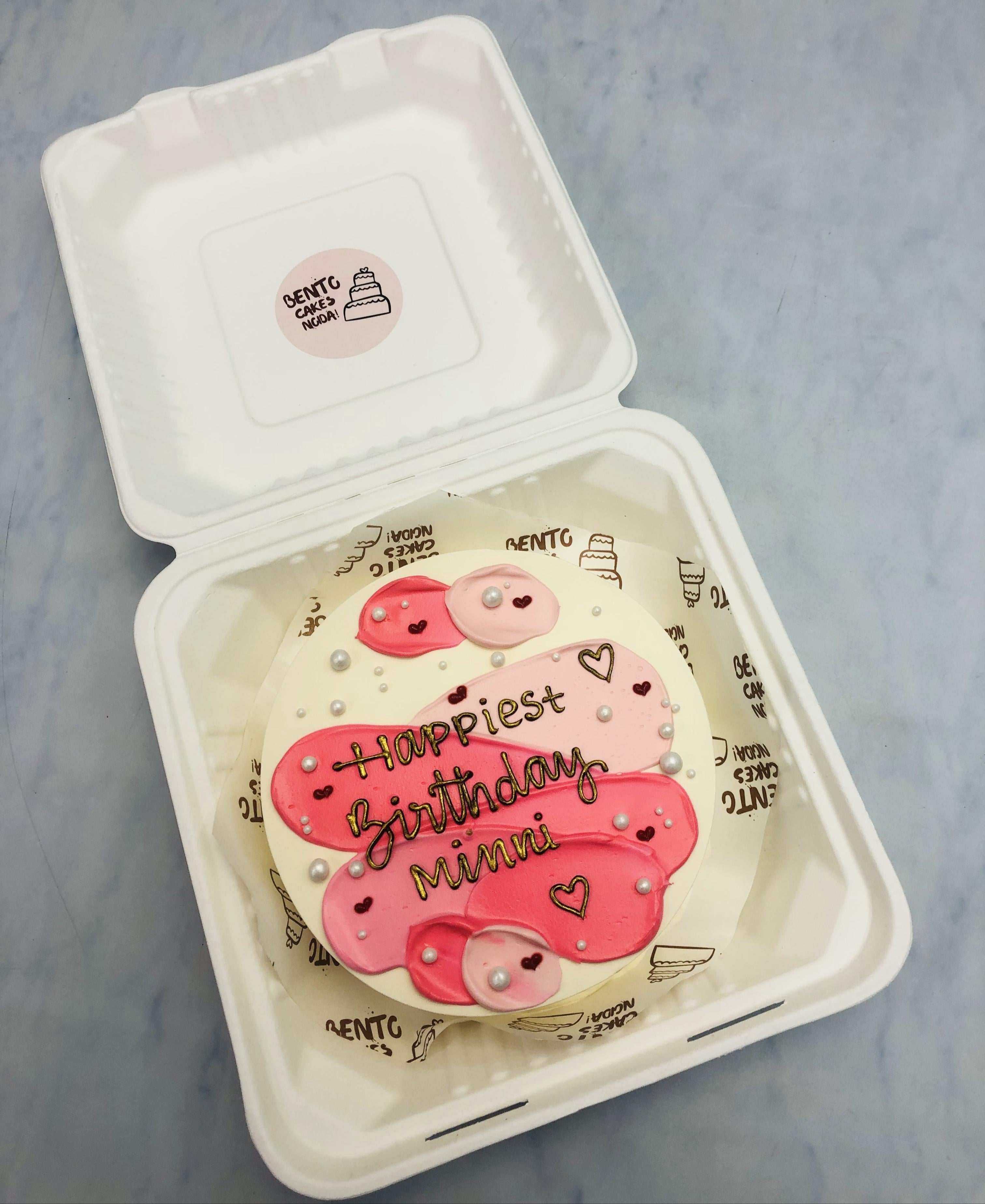 A small round birthday cake with pink and white icing, decorated with pearls and hearts, placed inside a white bento box. The cake has the message 'Happiest Birthday Minni' written on it."