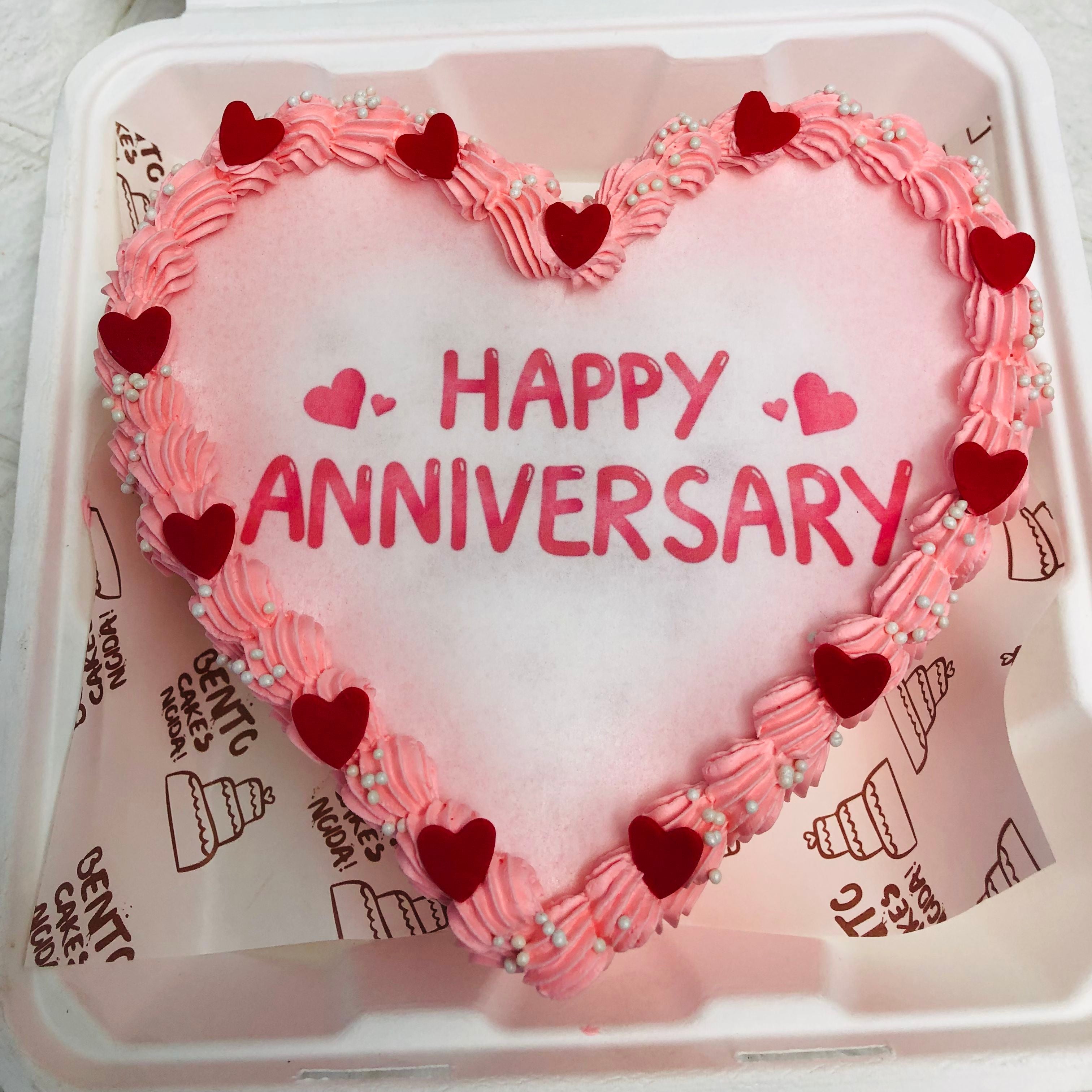 A delightful heart-shaped cake featuring "Happy Anniversary" written on top visible from front angle, ideal for a romantic celebration. 