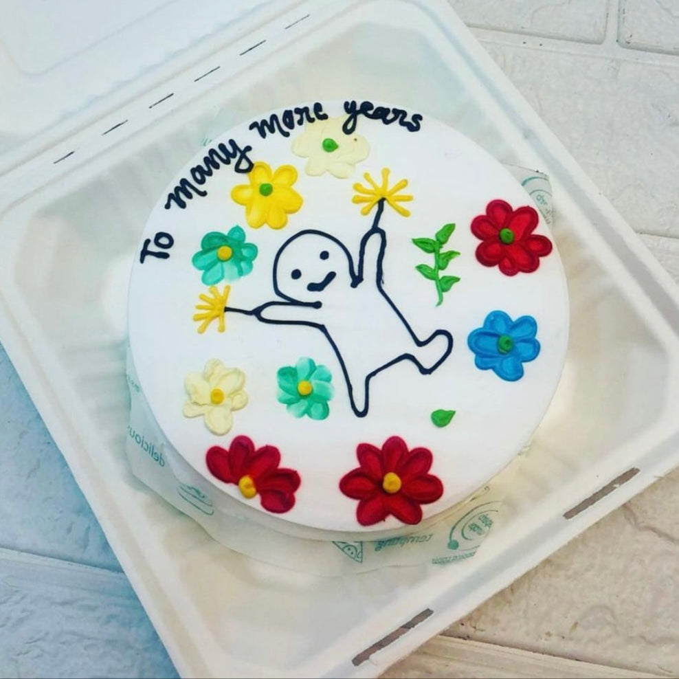 A round white cake with colorful flower decorations and a simple stick figure drawing holding yellow flowers. The cake has the text "To many more years" written on it and is placed inside a bento box.