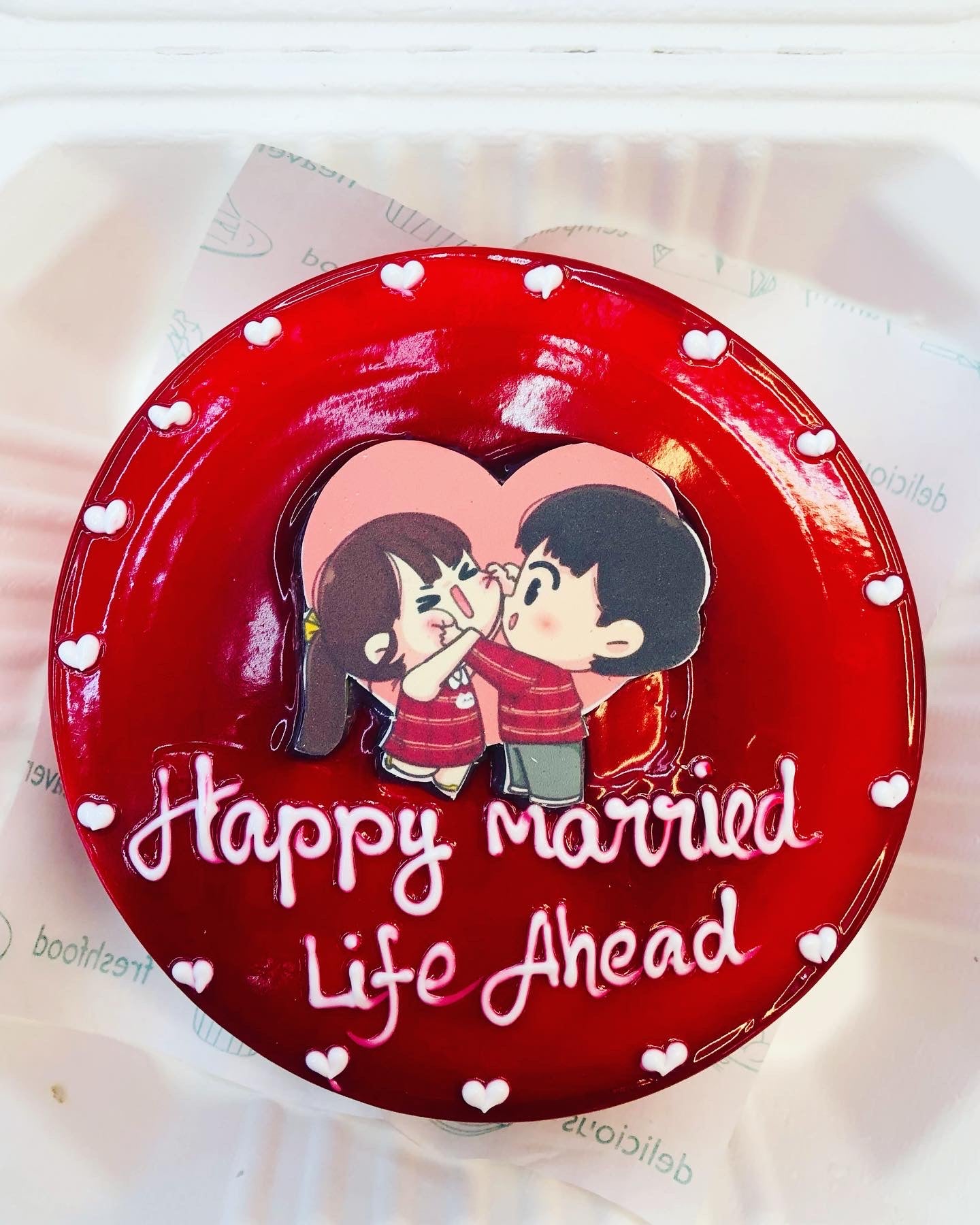 A Jelly Red cake designed with little white hearts and a cute cartoon couple playing sticker placed on cake and a text "Happy Married Life Ahead" written with white icing.