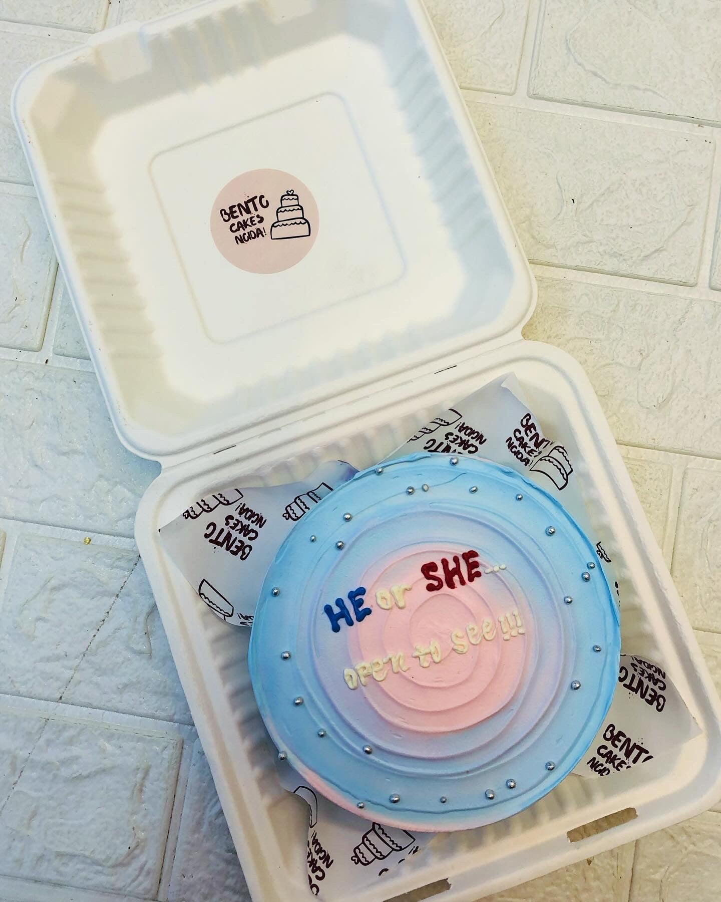 A blue pink ombry cake wit HE or SHE text on it and decorated with silver sprinkles.