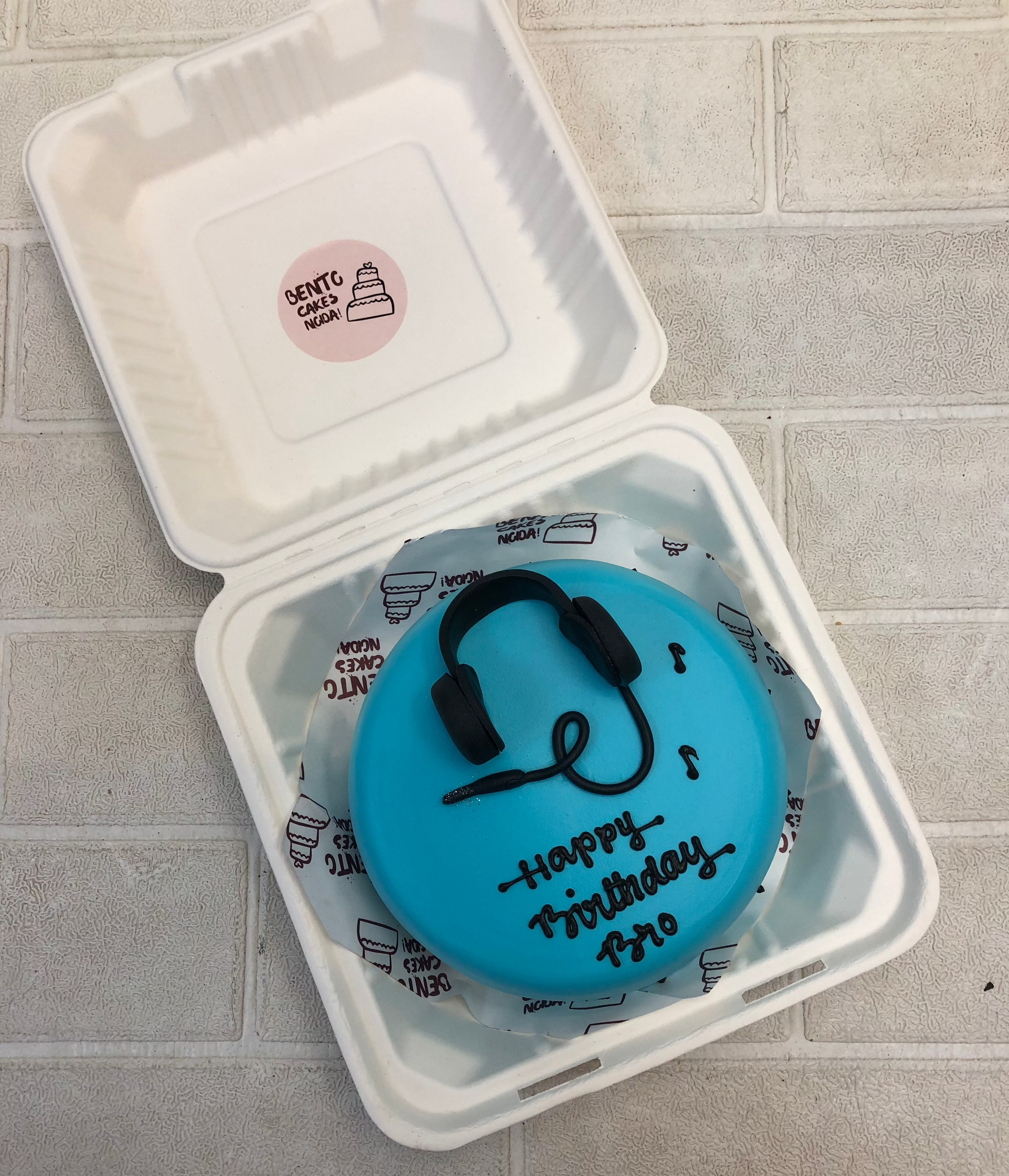 A vibrant blue birthday cake featuring a black headphone design with musical notes. The cake has "Happy Birthday Bro" inscribed on top, making it a perfect treat for a music lover.