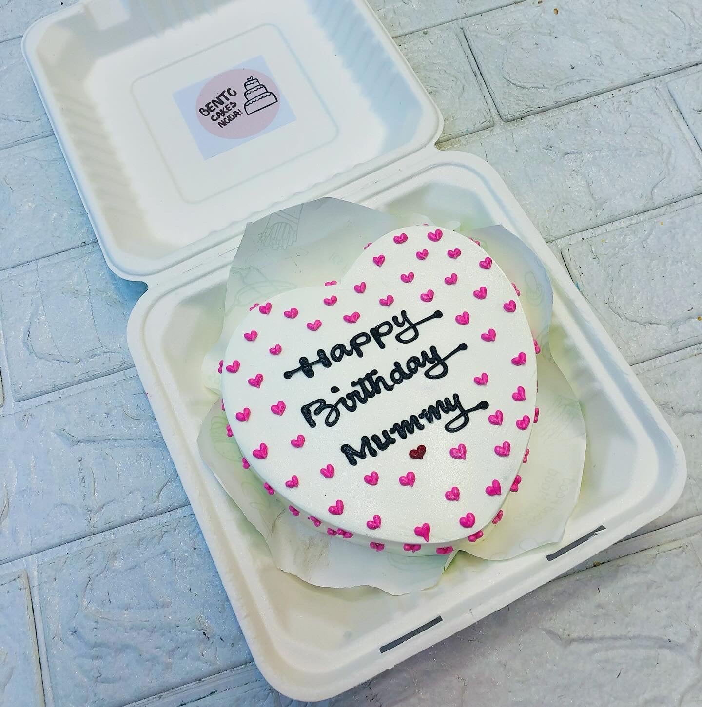 A heart-shaped birthday cake with white icing, decorated with small pink hearts and featuring the message "Happy Birthday Mummy" written in black icing. 
