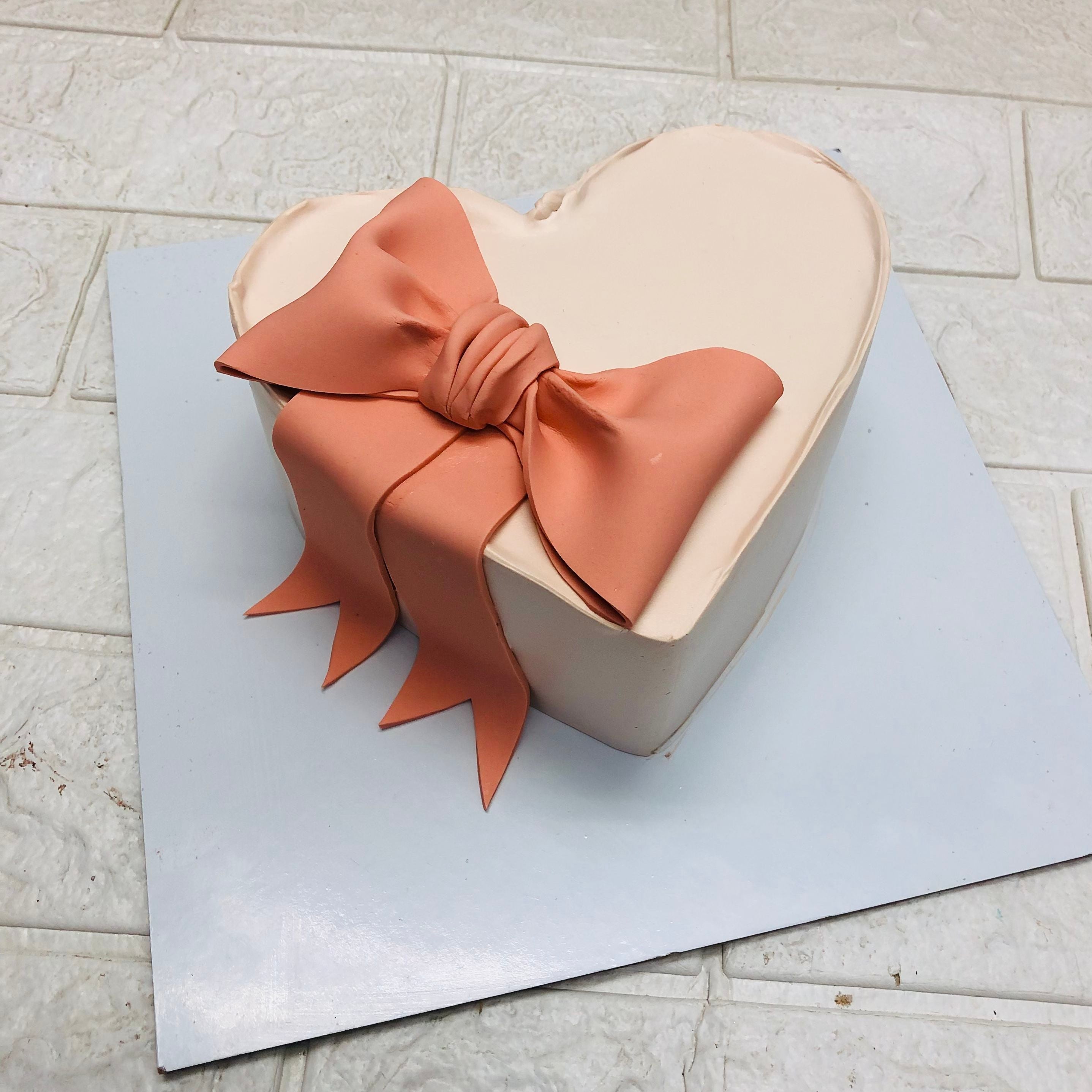A heart-shaped cake covered in smooth white fondant, adorned with a large, elegant peach-colored bow on top, placed on a white square board.