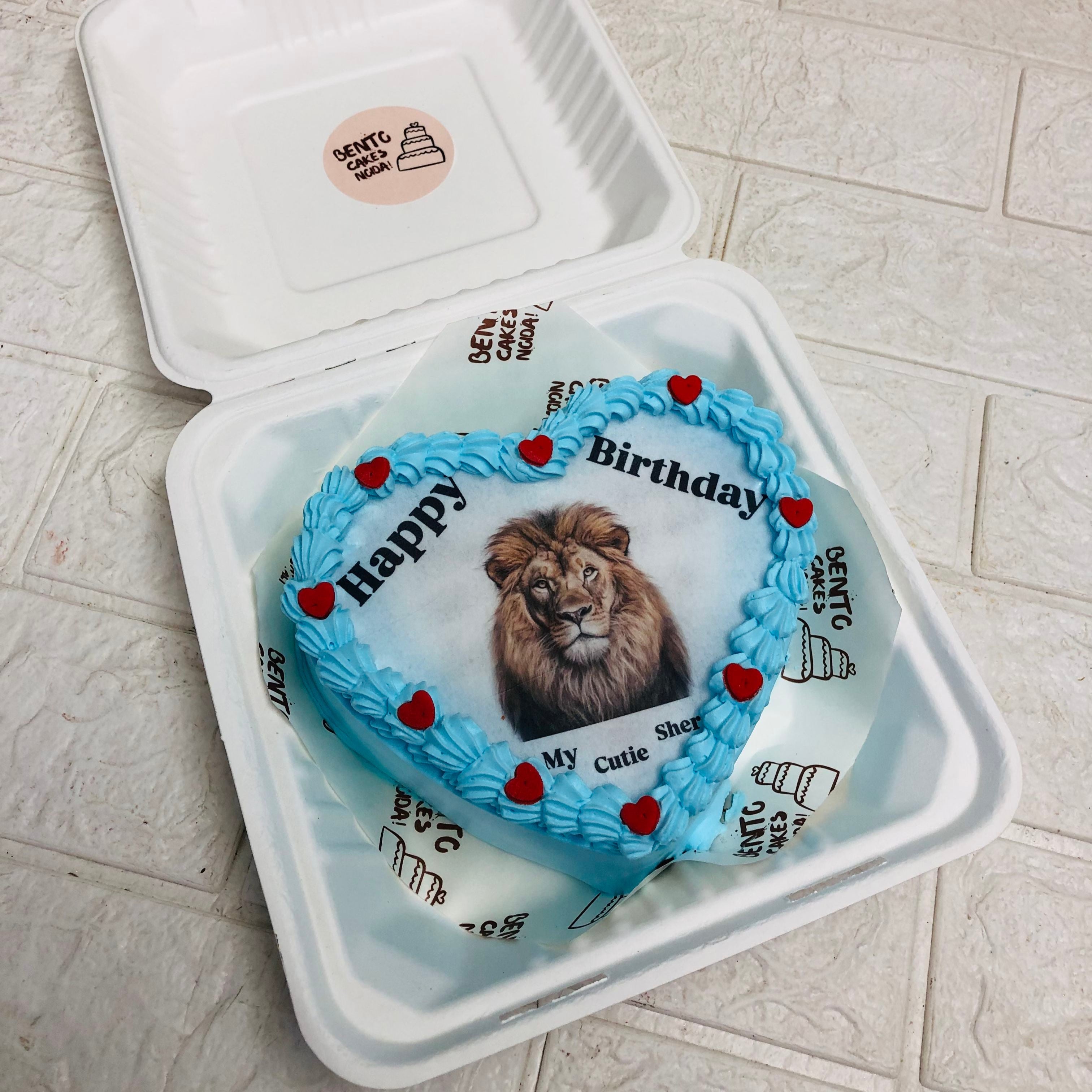 A heart-shaped birthday cake with blue frosting, decorated with red heart-shaped candies, and featuring an image of a lion in the center.