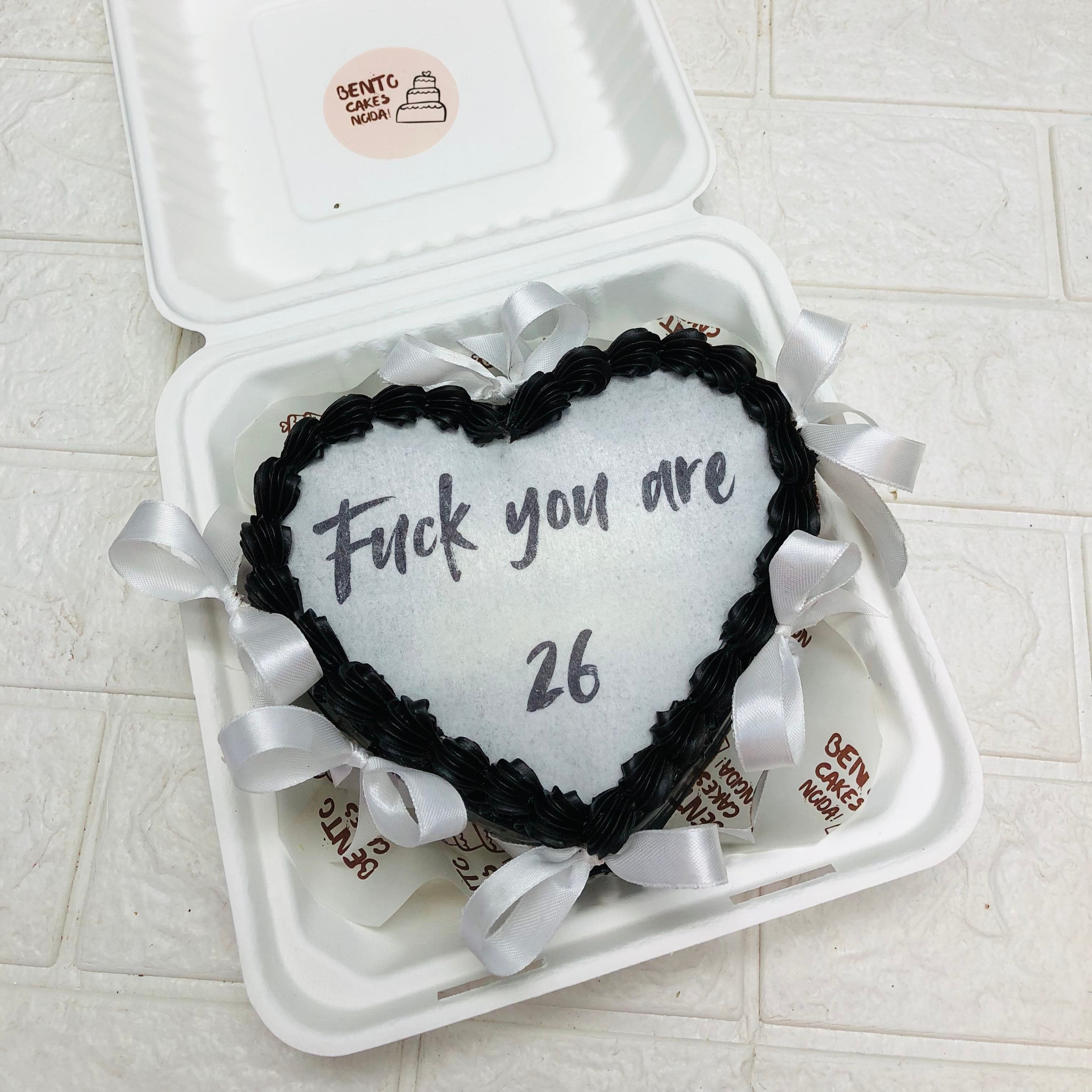 A heart-shaped cake with black icing and white ribbons, placed inside a white takeout box. The cake has a message written in black icing that reads, "Fuck you are 26."