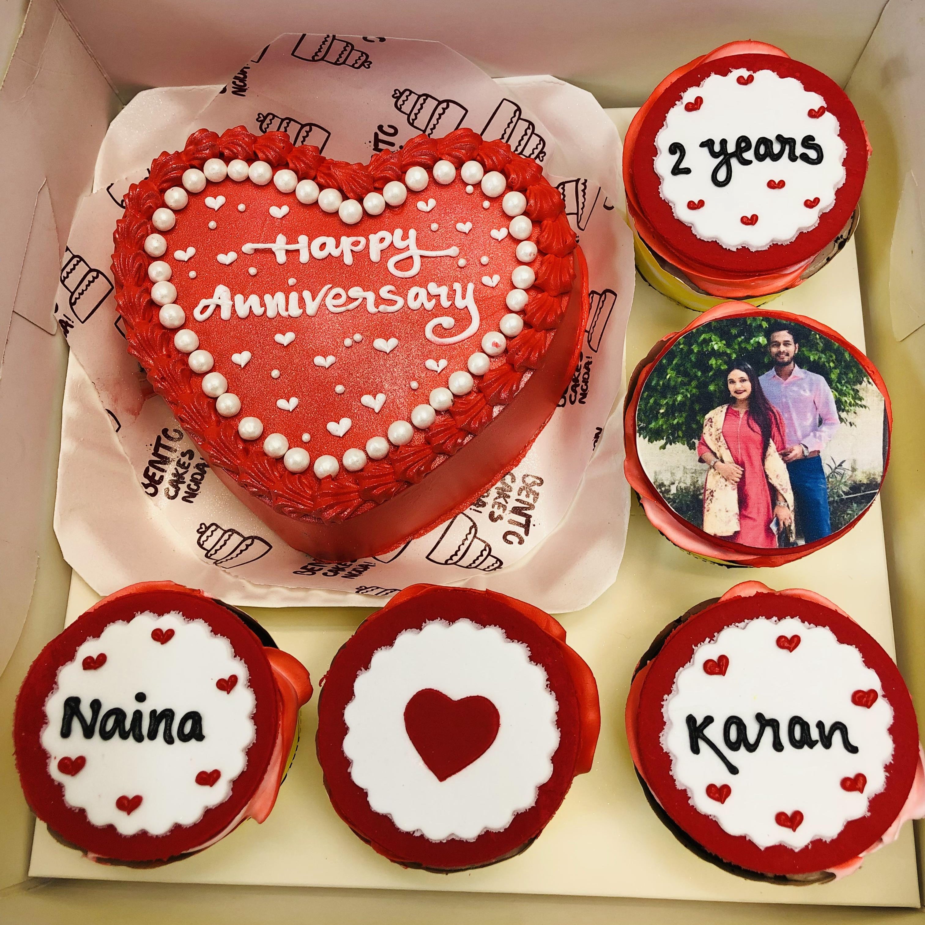 A heart-shaped red anniversary cake with "Happy Anniversary" written on it in white icing, surrounded by four cupcakes. The cupcakes display designs featuring "Naina," "Karan," a red heart, "2 years," and a photo of a couple.