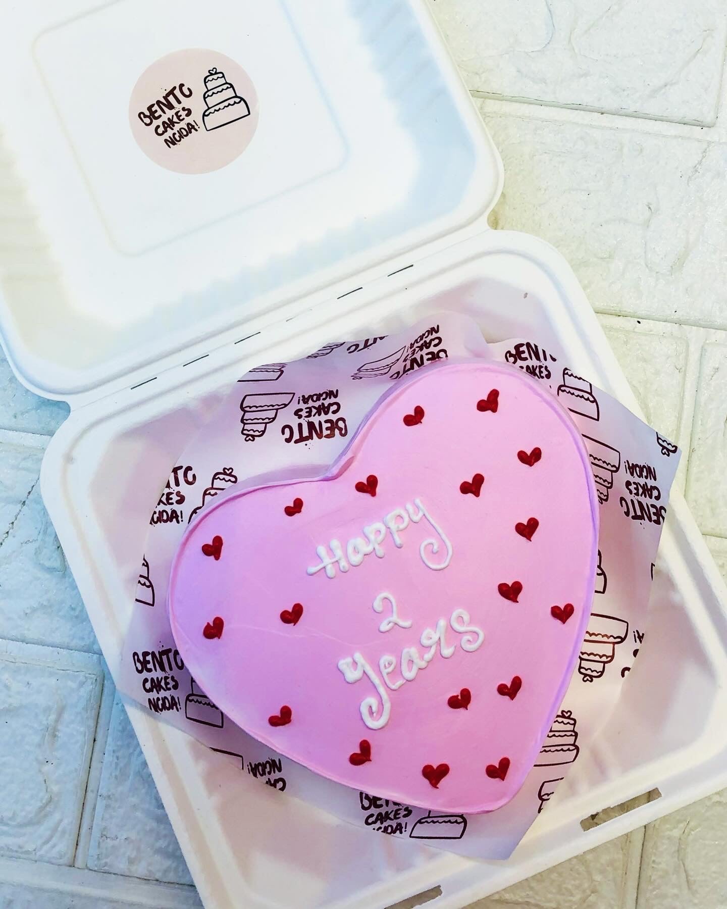 A light pink heart shape cake filled with red hearts design and a text written with white icing on it.