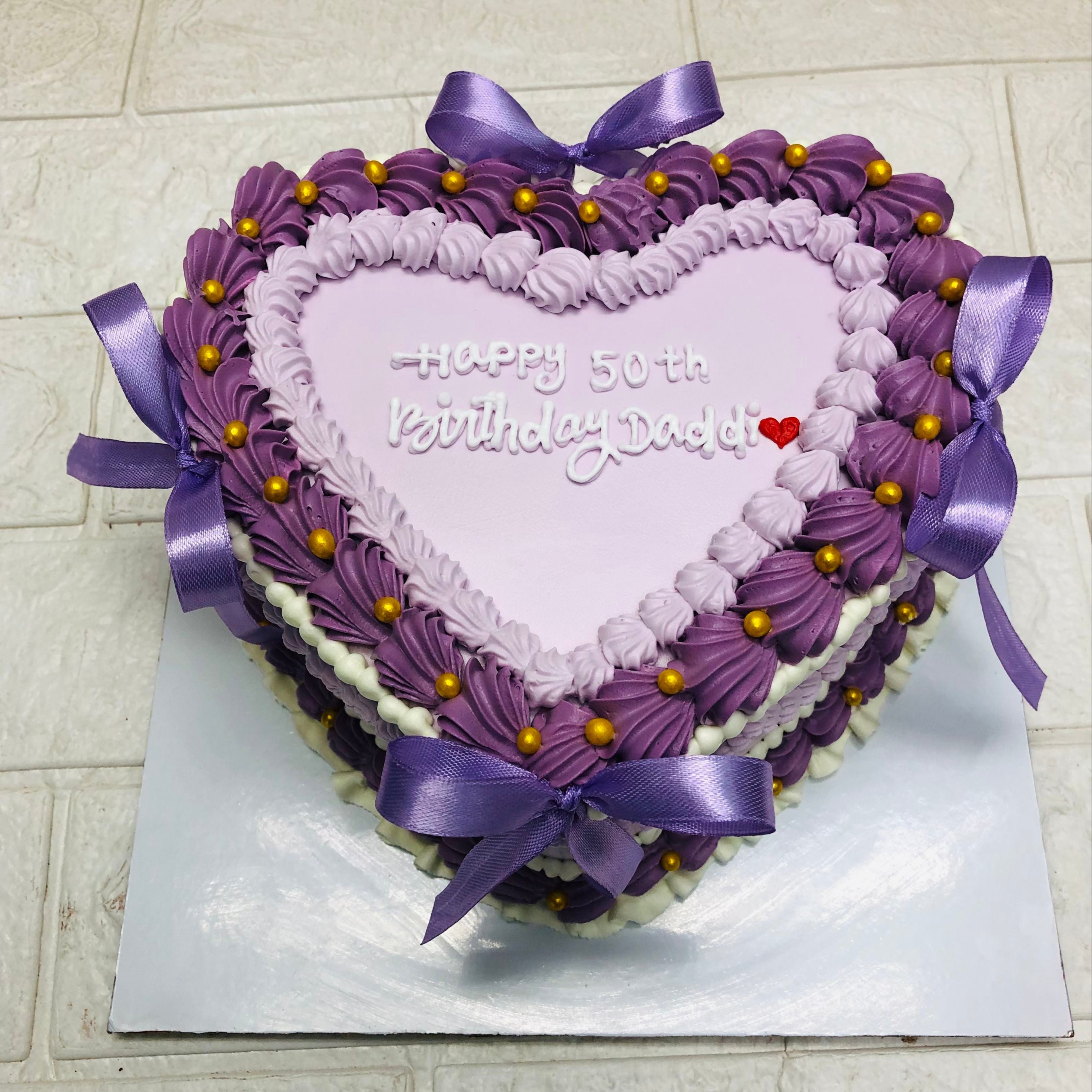 A heart-shaped cake decorated with purple and white frosting, purple ribbons, and golden beads, with the message "Happy 50th Birthday Dad!" written on top.
