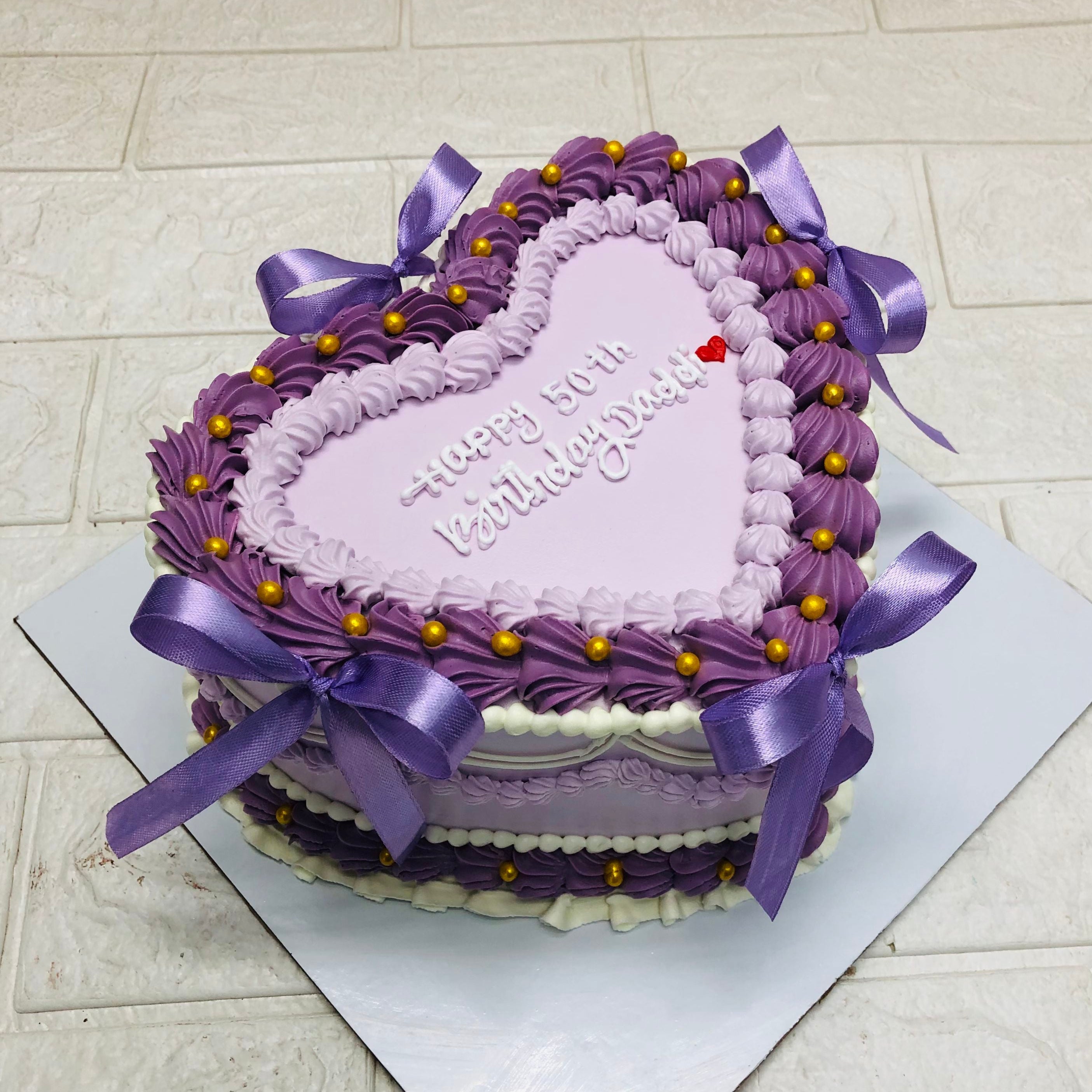 A heart-shaped cake decorated with purple and white frosting, purple ribbons, and golden beads, with the message "Happy 50th Birthday Dad!" written on top.