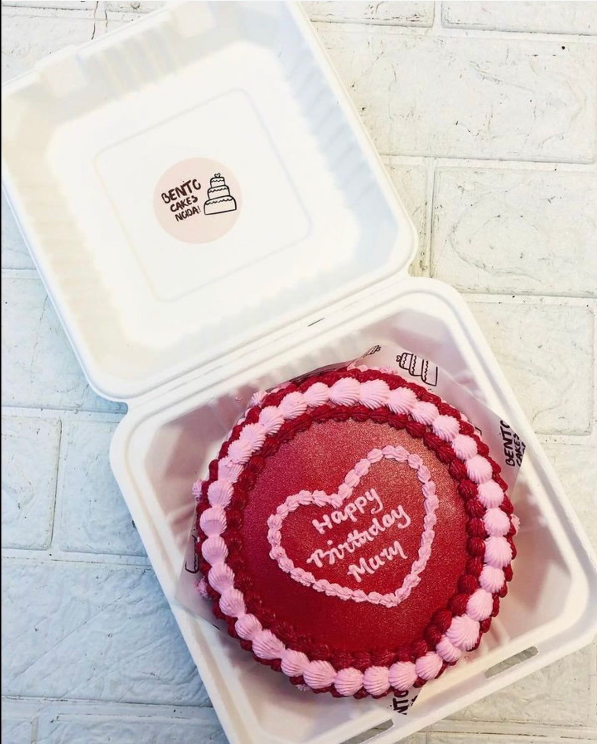 A heart-shaped red velvet birthday cake with pink and red frosting, inscribed with 'Happy Birthday Mum,' placed inside a bento box.