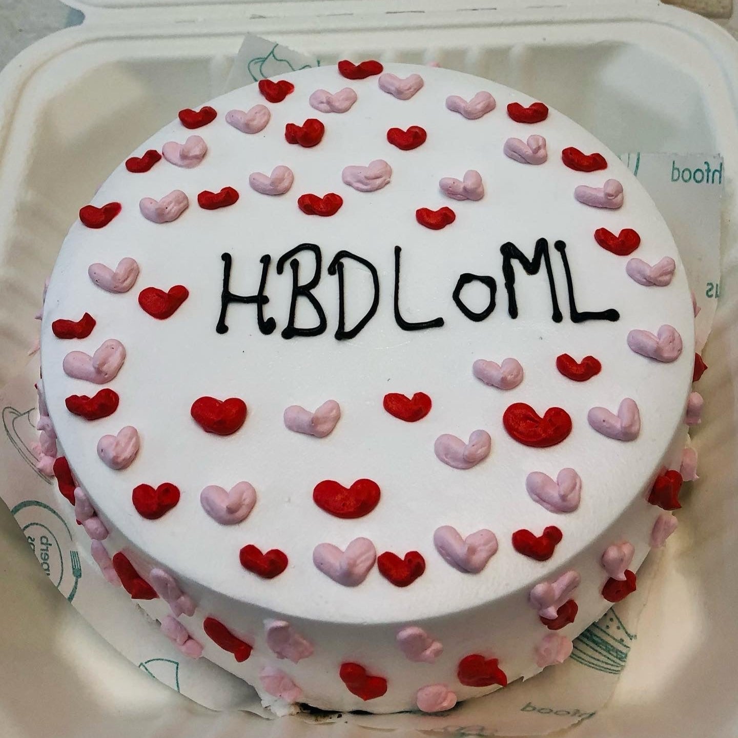 A round white cake decorated with red and pink heart-shaped icing, featuring the text 'HBDLoML' written in black icing on the top.