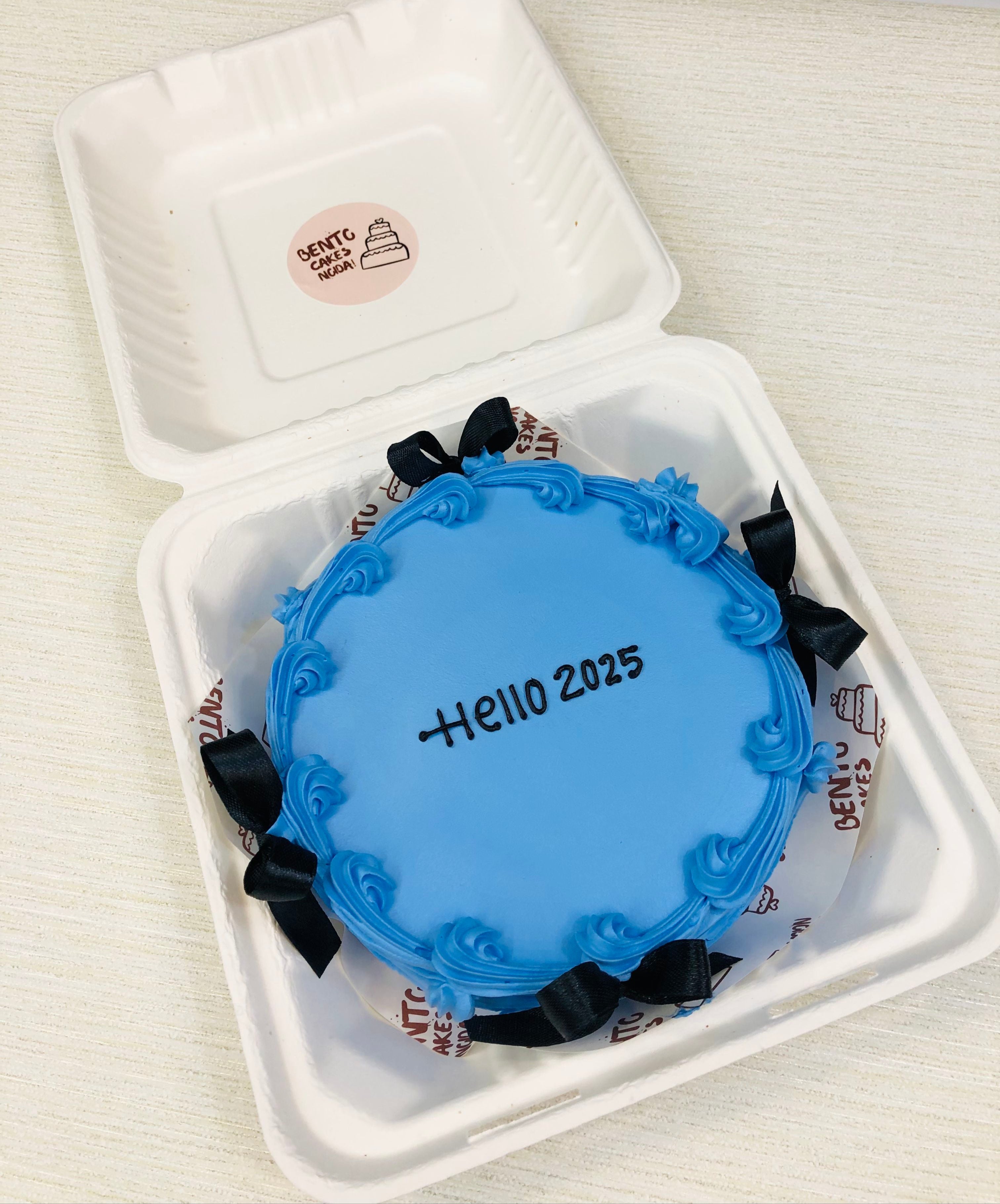 A blue frosting bento cake decorated with black bows and a text "Hello 2025" written with white icing.