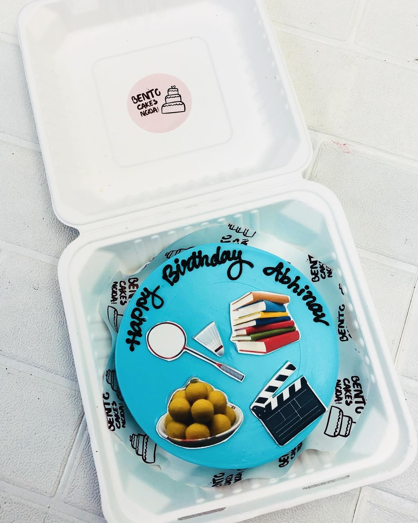 A light blue bento cake is designd for bitrthday having his hobbies printed on it.