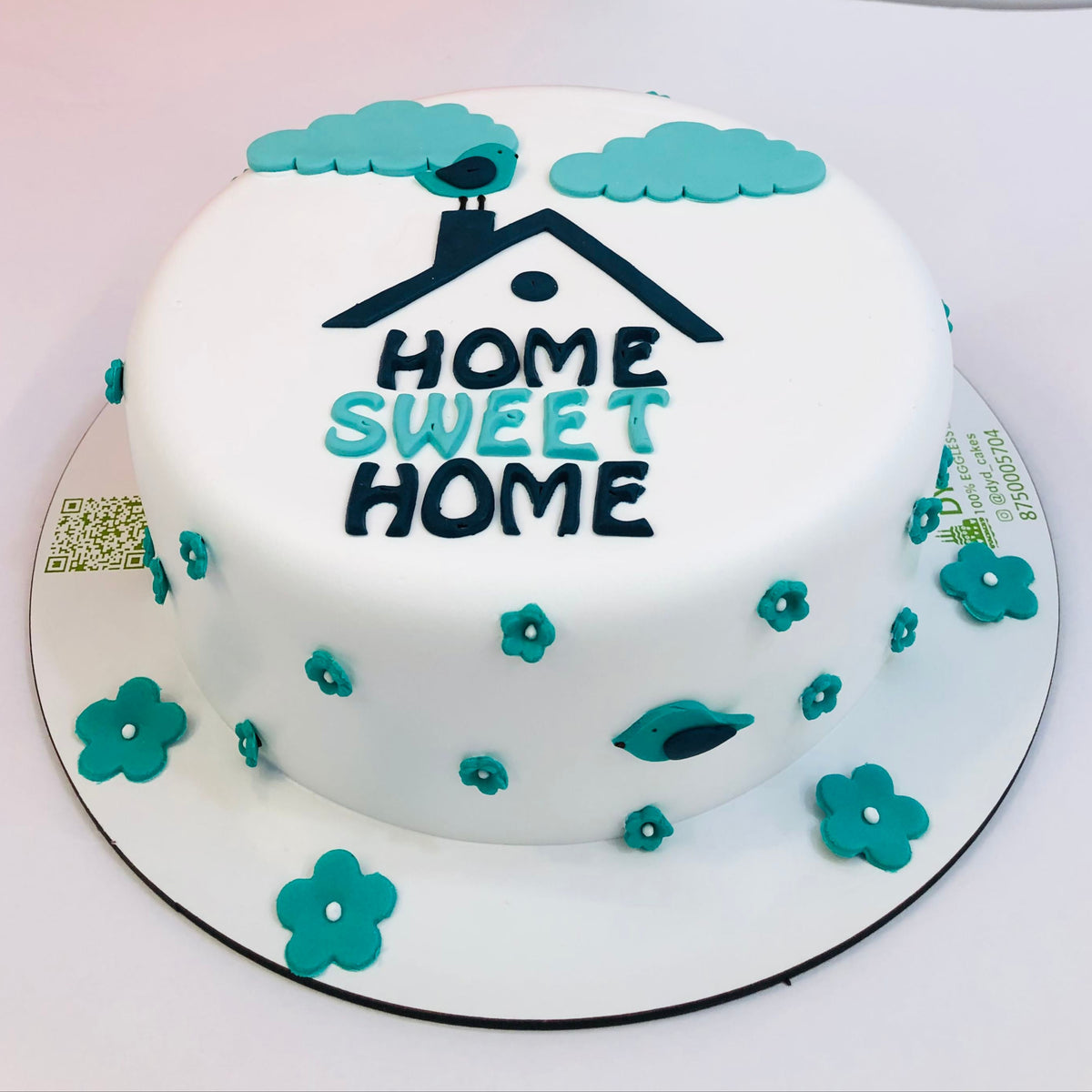 A round fondant cake with a 'Home Sweet Home' theme, decorated with teal and black fondant designs including a house, bird, clouds, and flowers.