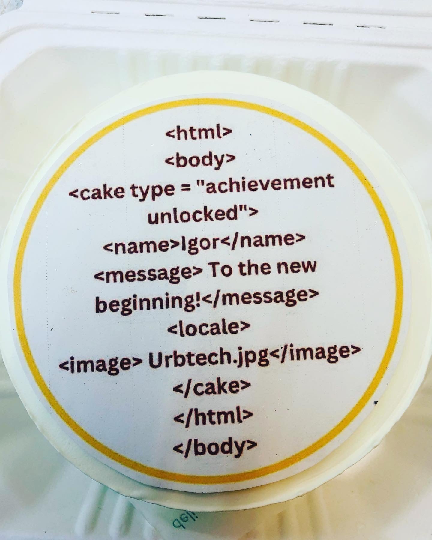 A round cake with a white icing top, featuring HTML-like text in brown. The text includes tags such as <html>, <body>, <cake type="achievement unlocked">, etc. The cake is bordered with a thin yellow line.