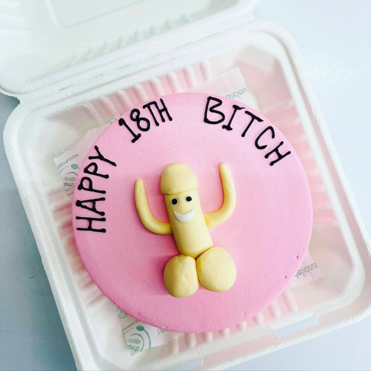 Pink cake with a humorous design featuring a smiling, anthropomorphic figure resembling male genitalia, with the text "HAPPY 18TH BITCH" written on top, placed inside a white bento box.