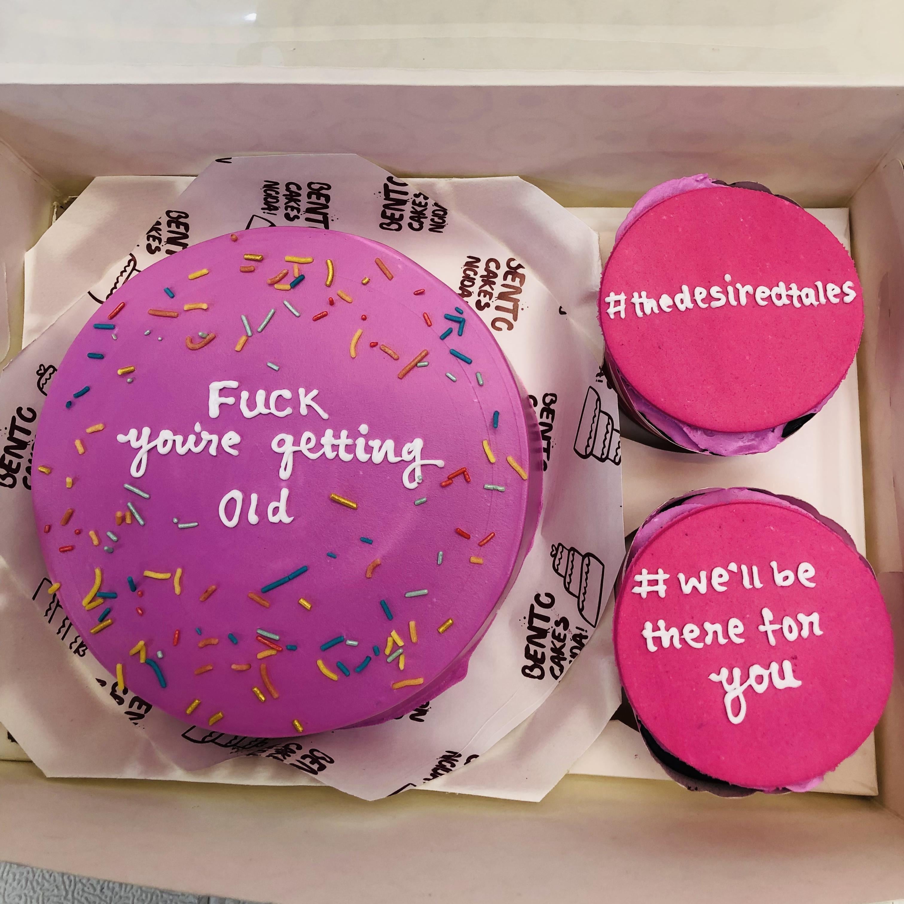 A set of three purple mini bento cakes in a box, decorated with colorful sprinkles. The largest cake reads "Fuck you're getting old," while the two smaller cakes display "#thedesiredtales" and "#we'll be there for you."