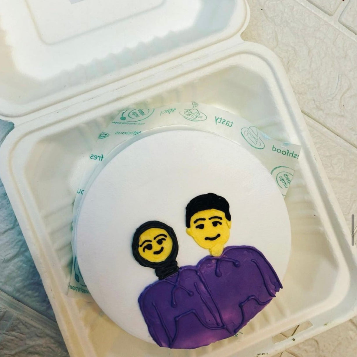 A round cake with a white frosting base, featuring a simple illustration of two people wearing purple hoodies. The cake is placed inside a bento box. One person is depicted with a yellow face and a black headscarf, while the other person's face is blurred.