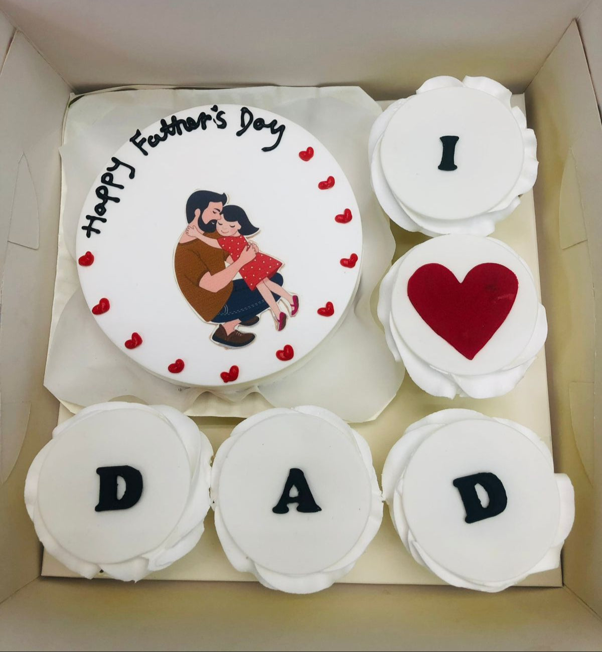 A white bento cake having fathers day theme of a father and daughter print and 5 cupakes with different alphabets.