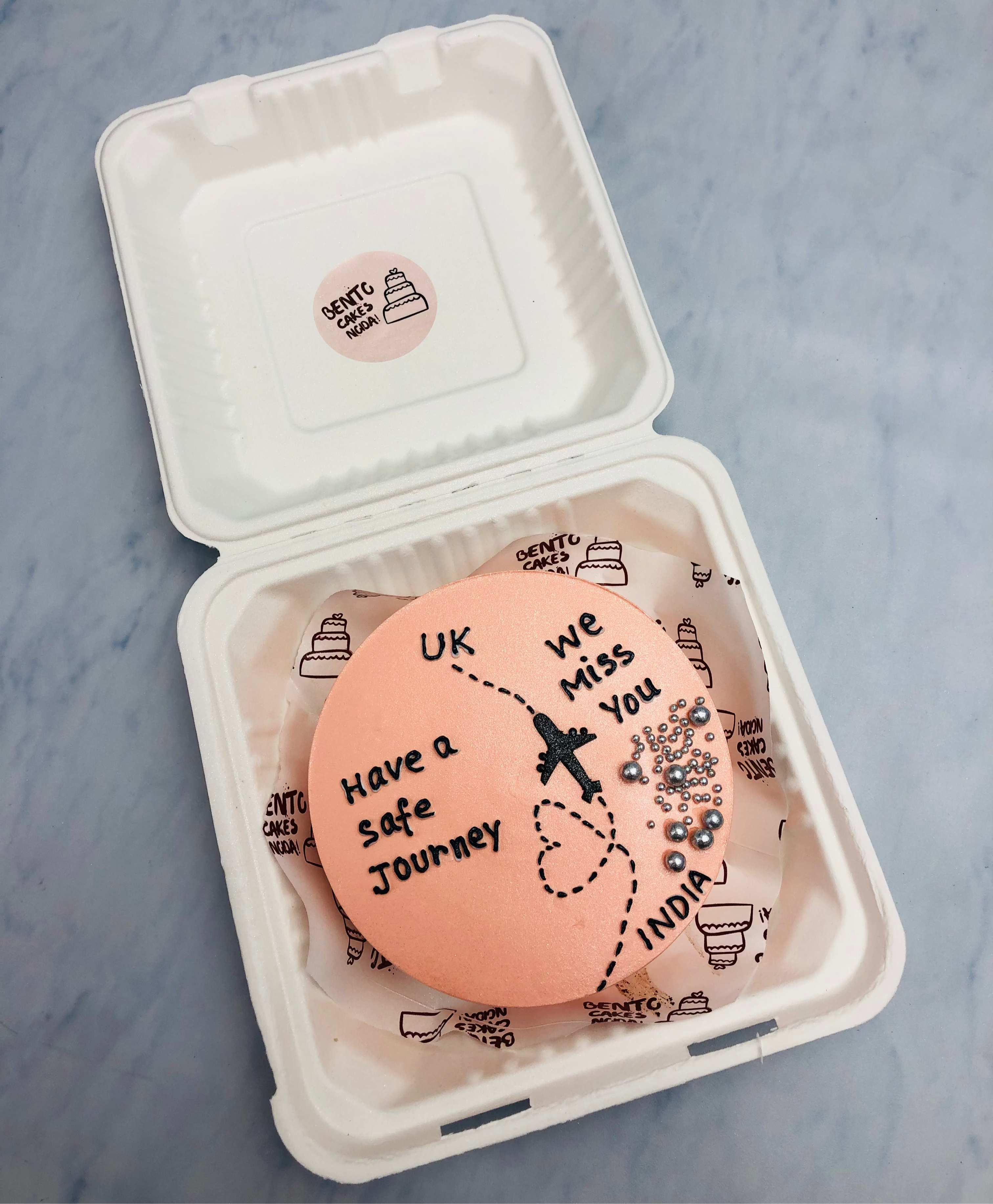 A peach color bento cake is designed for farewell theme and having a flight from India to UK with a safe journey message.