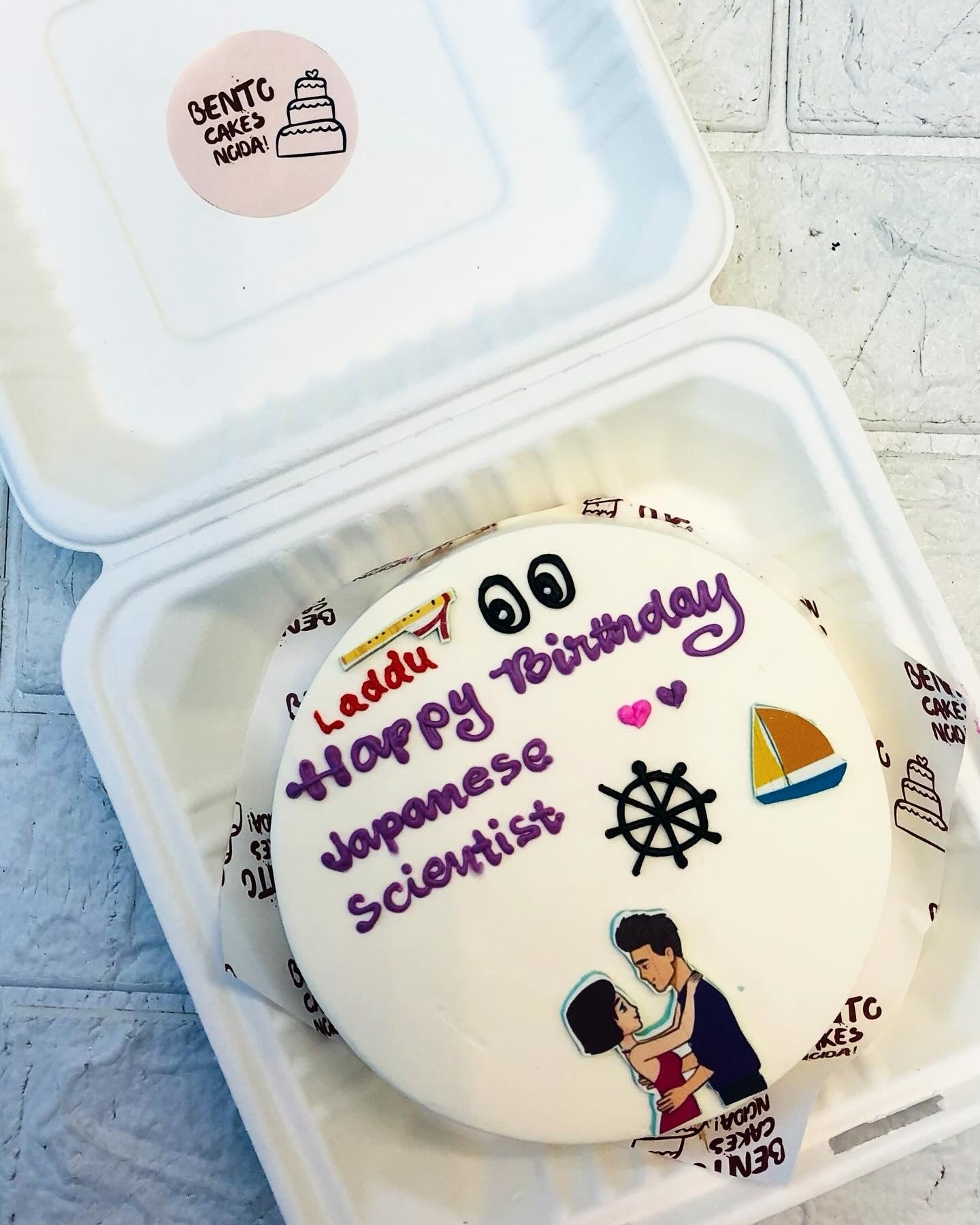 A white bento cake is designed with a loving couple sticker with a theme of Japanese Scientist for his birthday.