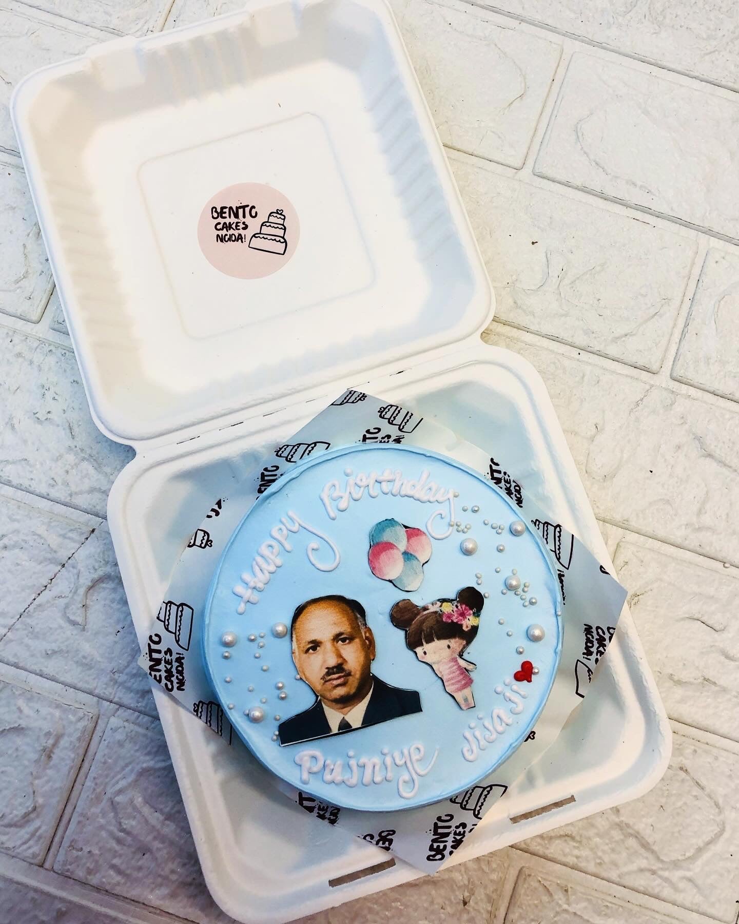 A light blue bento-style birthday cake with 'Happy Birthday Pujinye Jija ji' written in white icing. The cake is decorated with a cartoon girl, balloons, and pearl-like embellishments, placed inside a Bento Box.