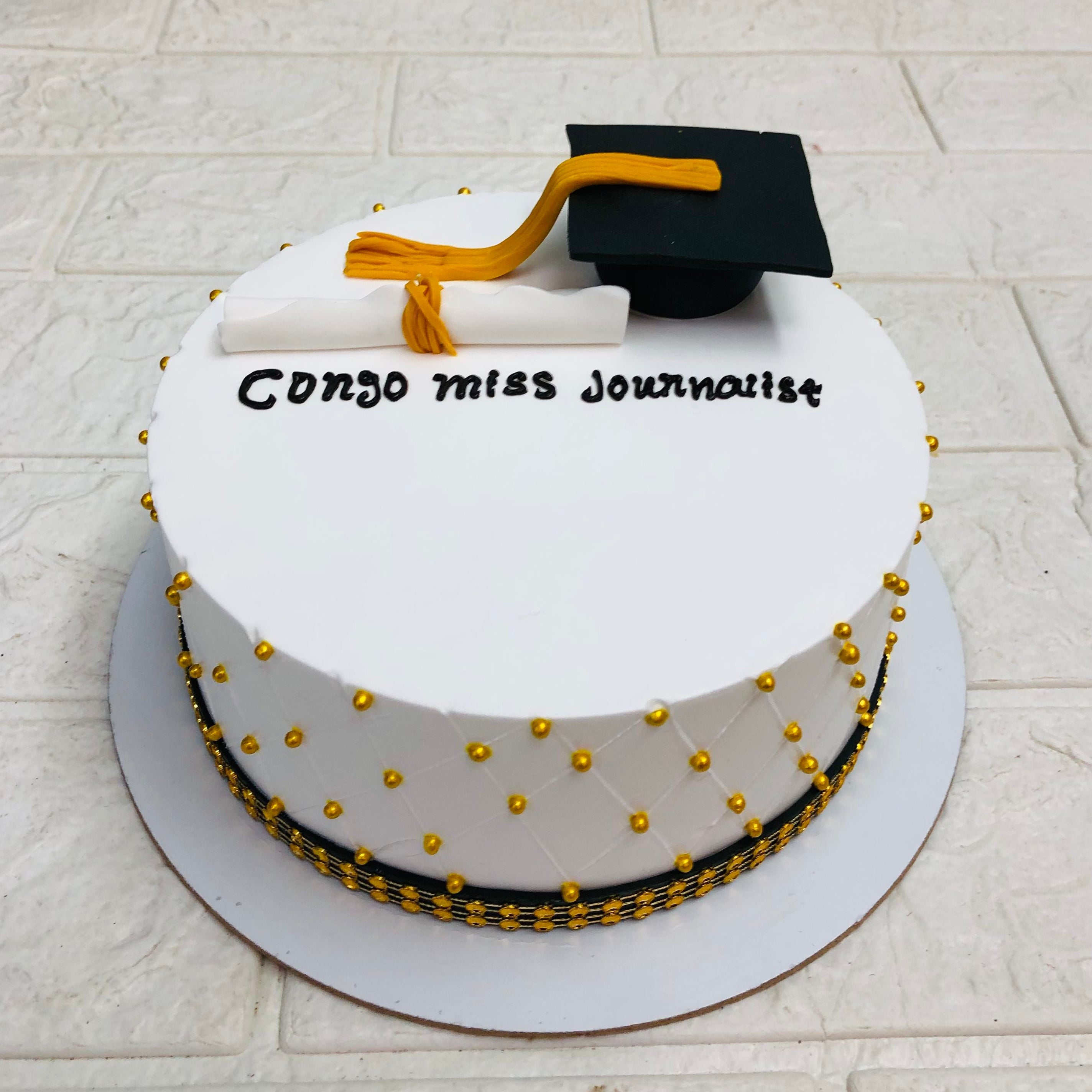 White graduation cake with gold decorations, topped with a black graduation cap and diploma, and the text "Congo miss Journalist" written in black icing.