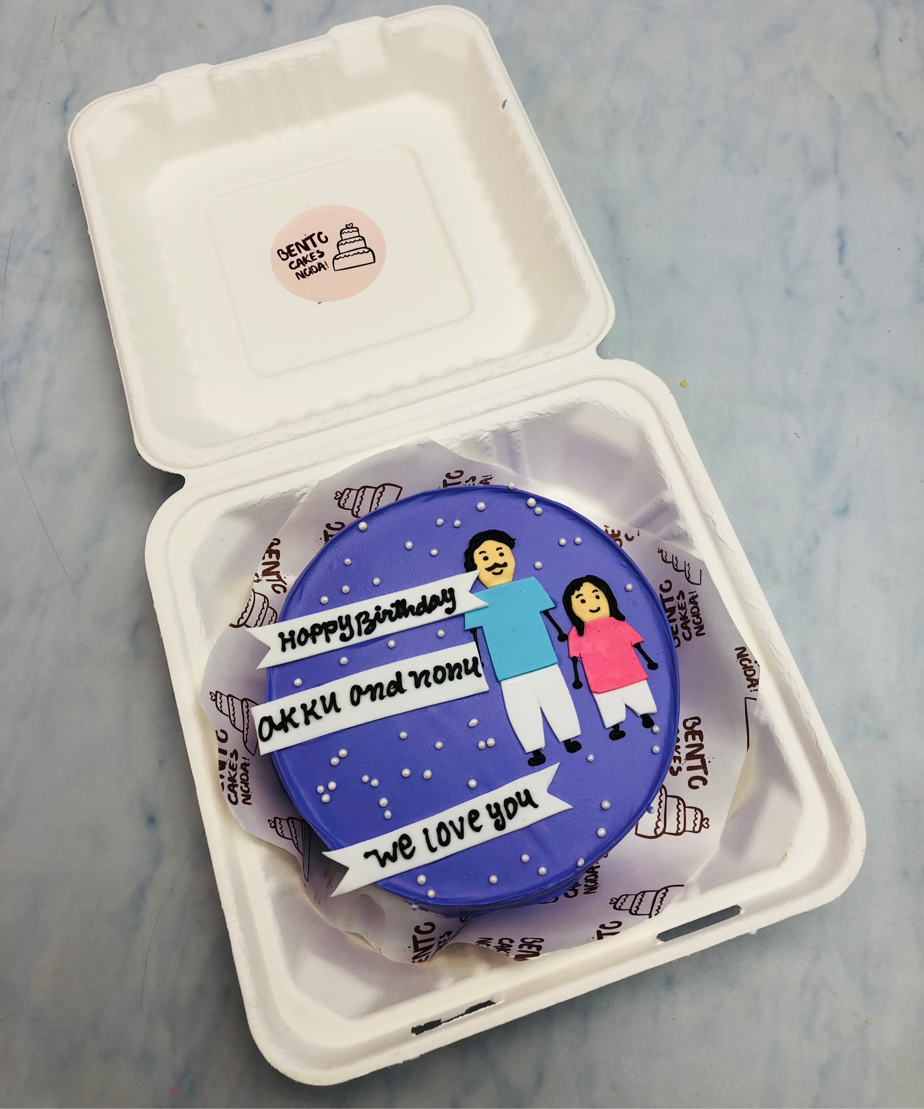 A custom birthday cake with blue frosting, featuring two figures holding hands and the text 'Happy Birthday Akku and Annu, We love you,' placed in a white bento box.