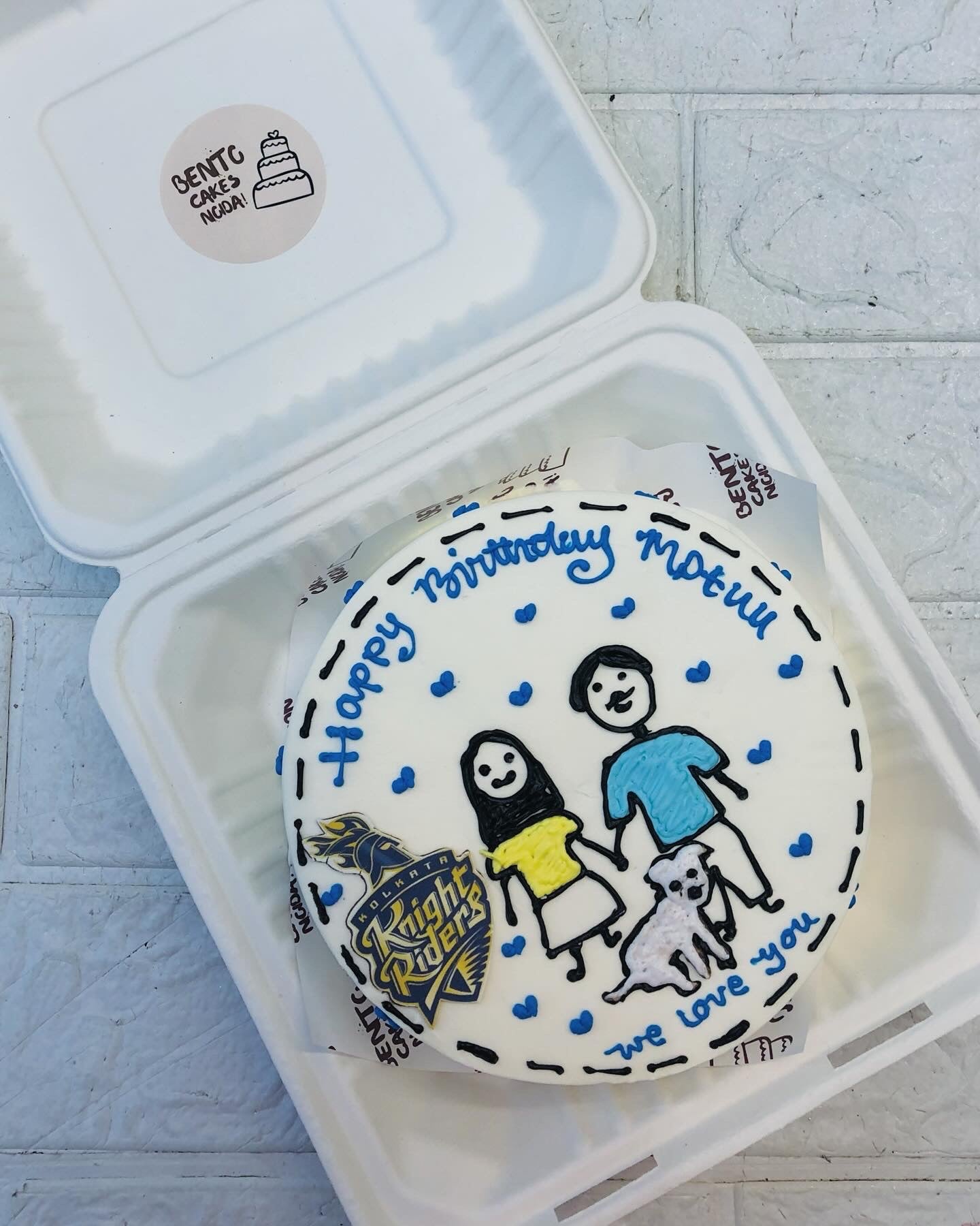 A custom-designed birthday cake inside a white box, featuring hand-drawn illustrations of two people and a dog, surrounded by blue hearts. The cake has the message "Happy Birthday Motu! We love you," along with a Knight Riders logo. 