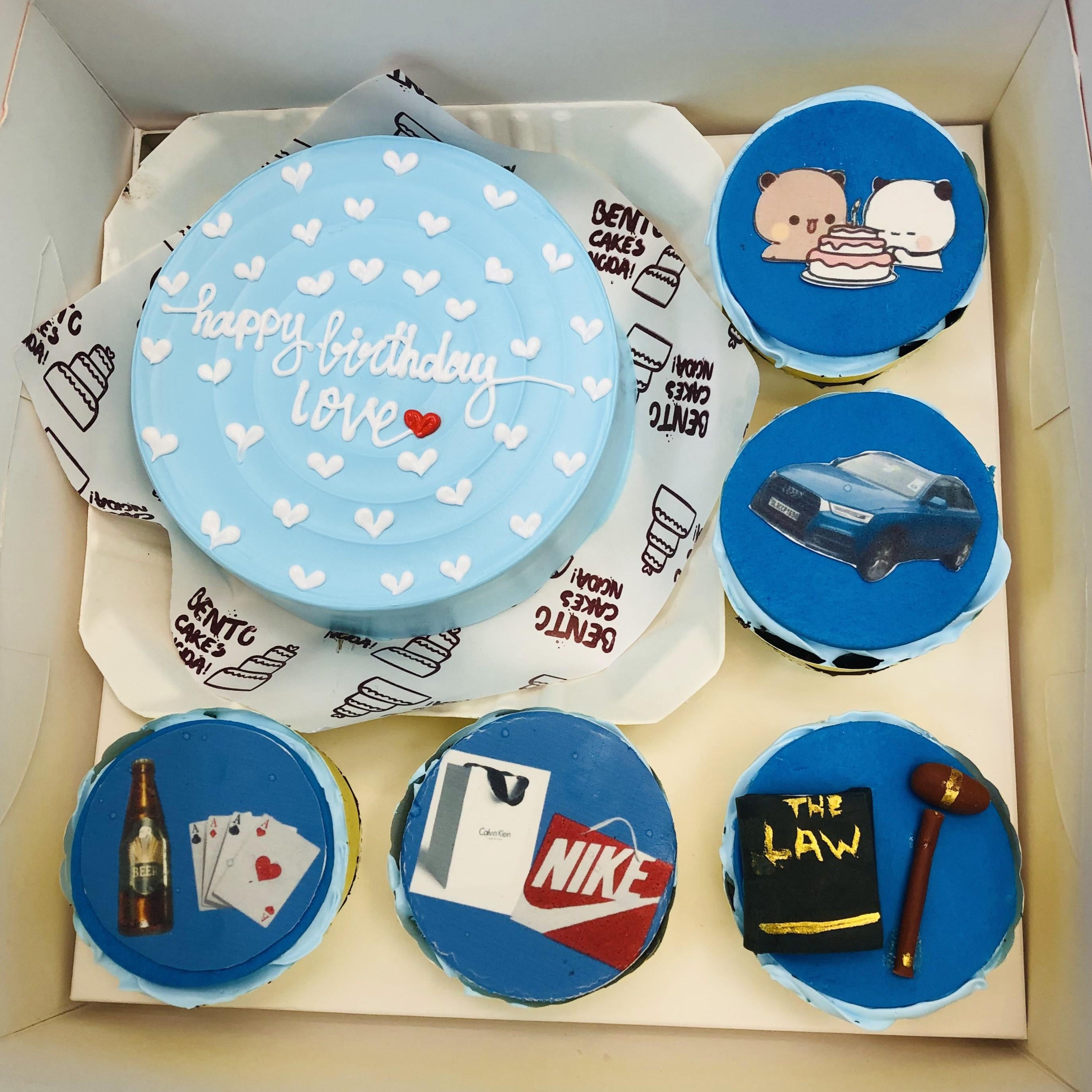 A lawyer-themed bento combo cake set with a central blue cake featuring the message 'Happy Birthday Love' and white hearts, surrounded by cupcakes with images including a law book and gavel, shopping bags, a beer bottle with playing cards, a blue car, and cartoon bears sharing a cake.