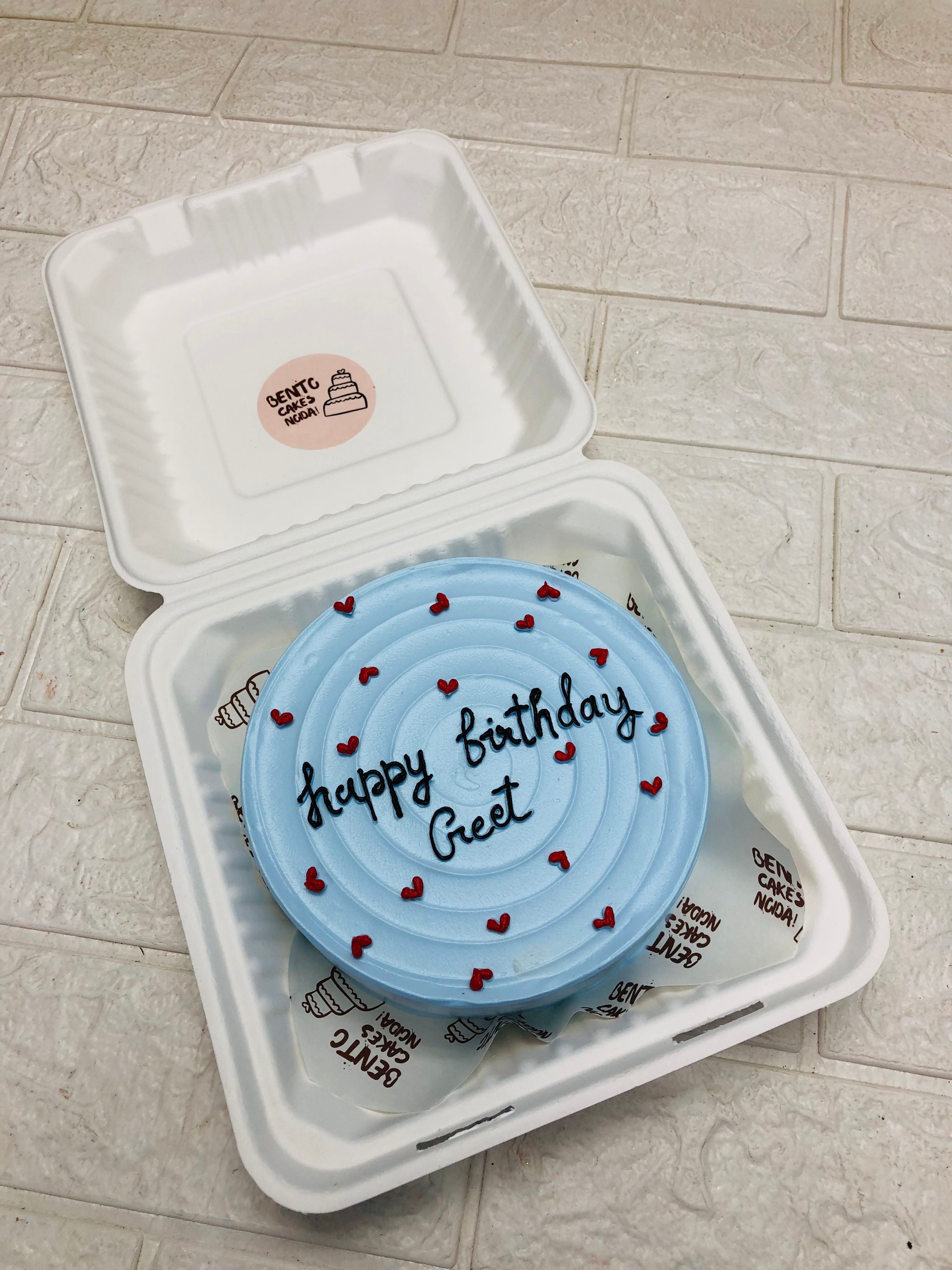 A light blue birthday cake with the message "Happy birthday Greet" written in black icing, decorated with small red hearts, placed inside a white biodegradable takeout box. The box has a sticker on the inside of the lid that reads "Bento Cakes Ncora."