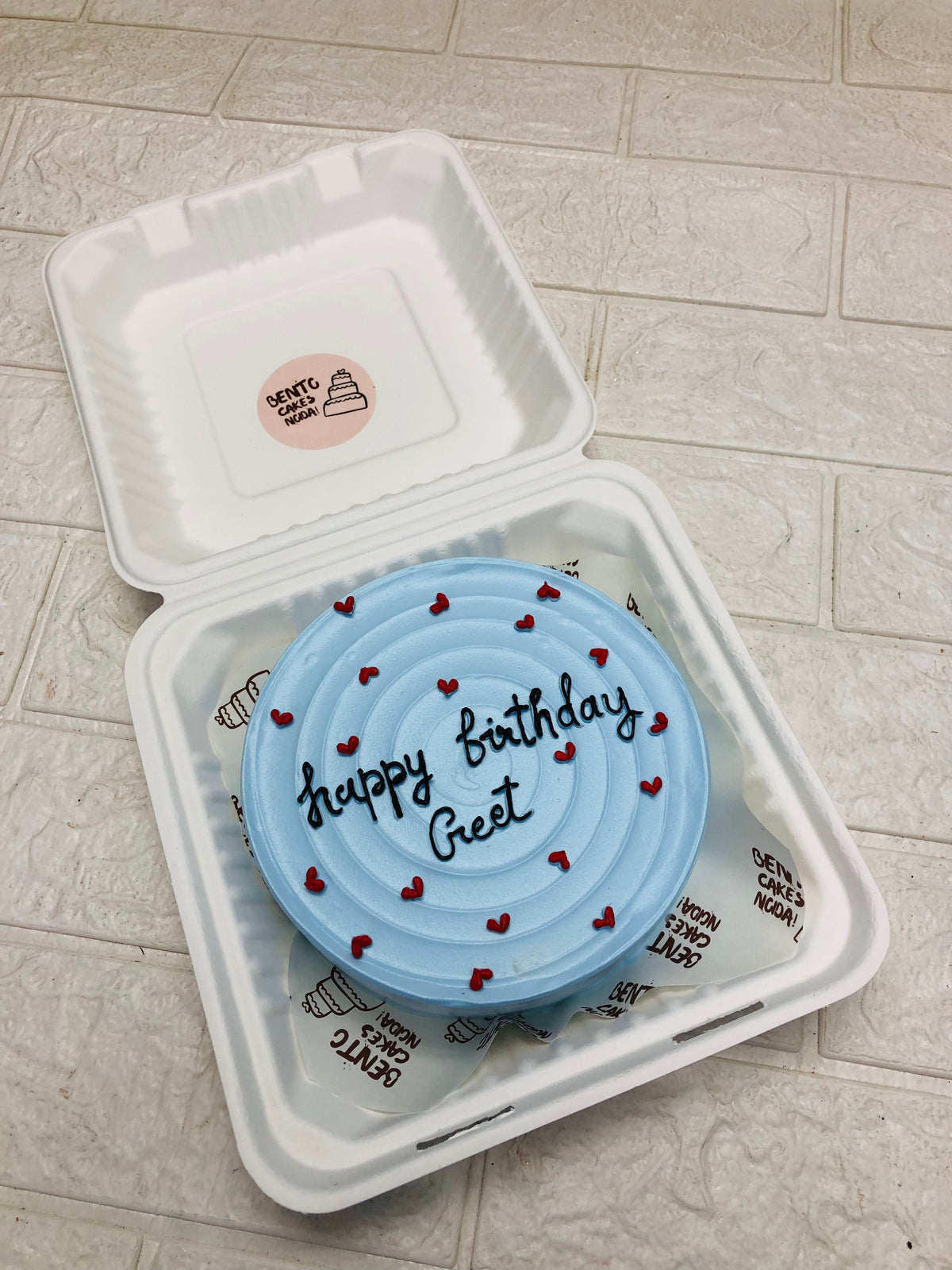 A light blue birthday cake with the message "Happy birthday Greet" written in black icing, decorated with small red hearts, placed inside a white biodegradable takeout box. The box has a sticker on the inside of the lid that reads "Bento Cakes Ncora."