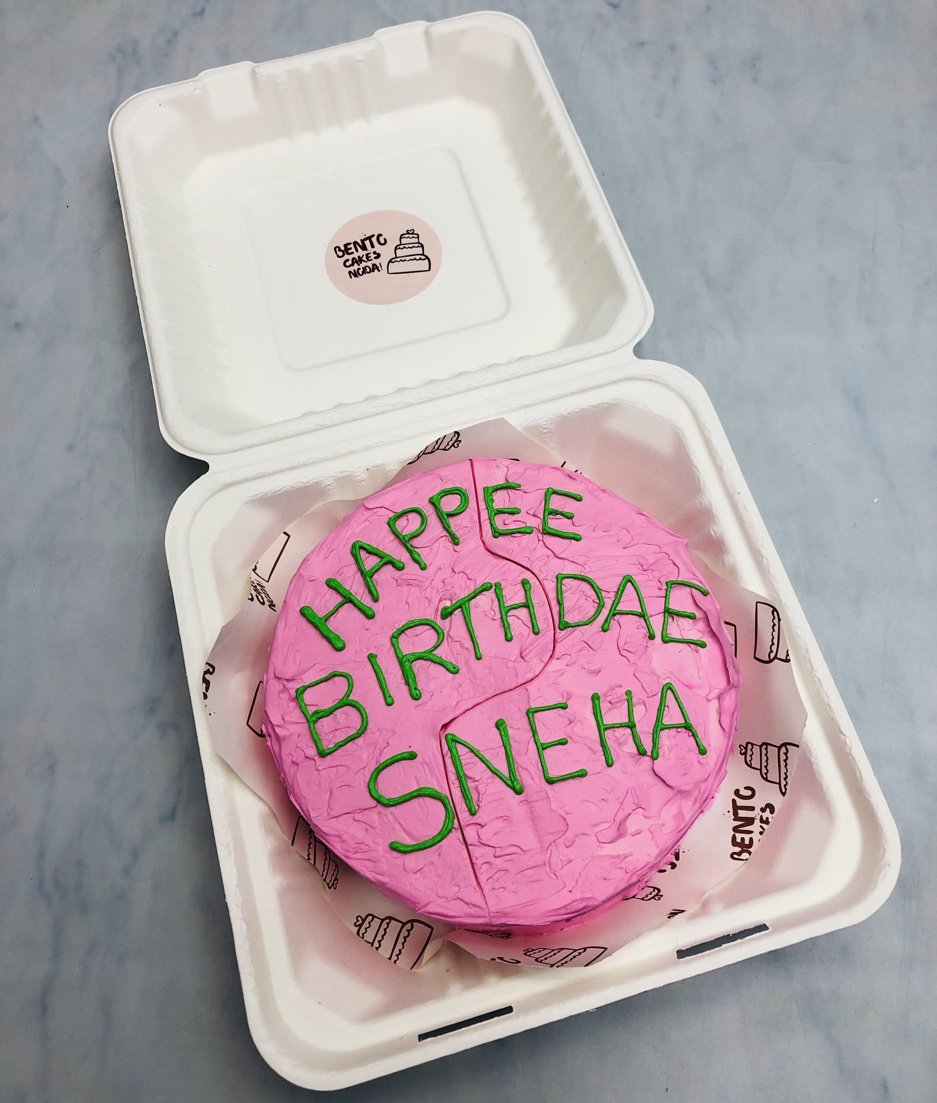 A pink birthday cake with the message 'HAPPEE BIRTHDAE SNEHA' written in green icing, placed inside a  bento box.