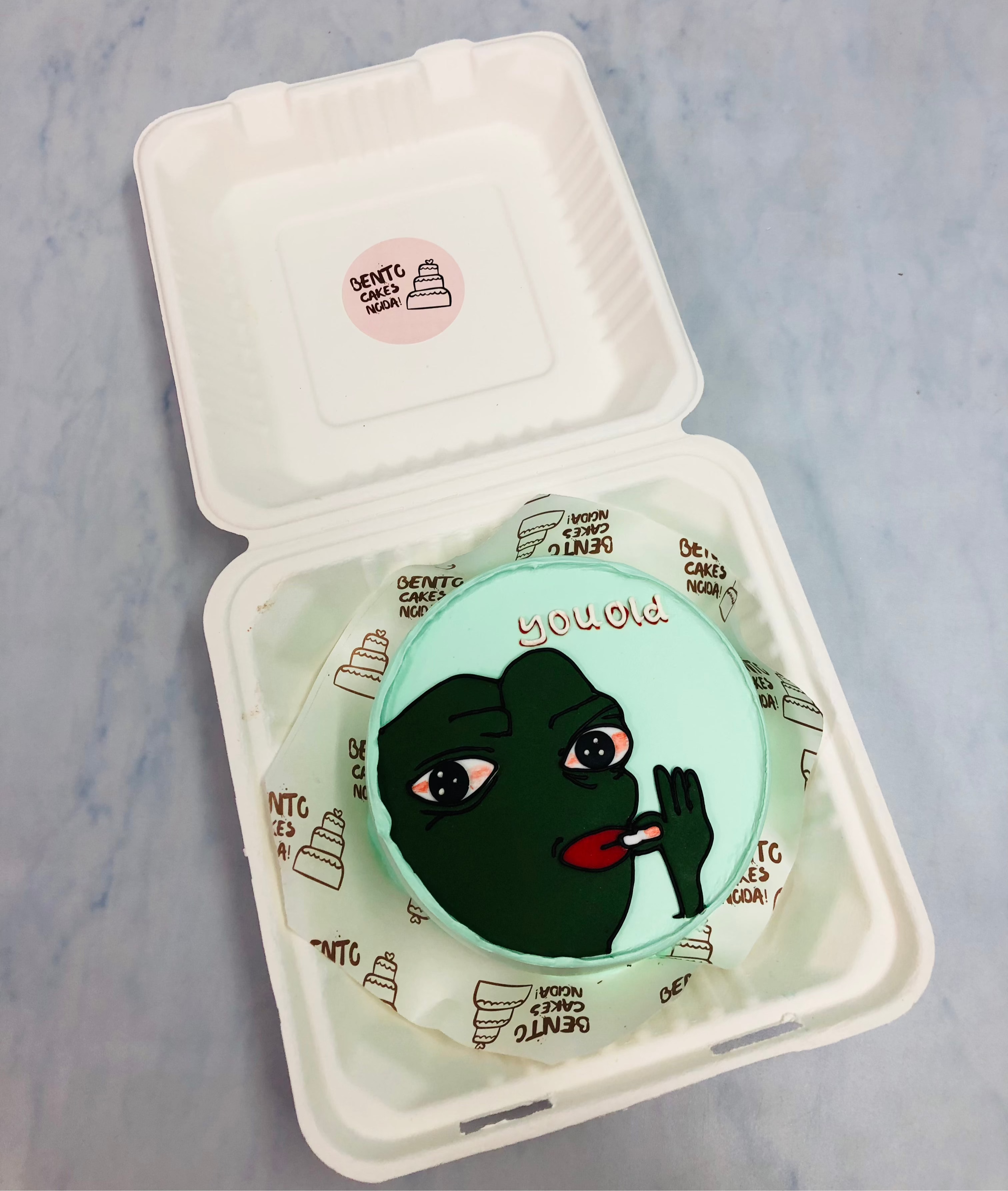 A lime blue bento cake is designed for smokers birthday having a green cartoon smoking.