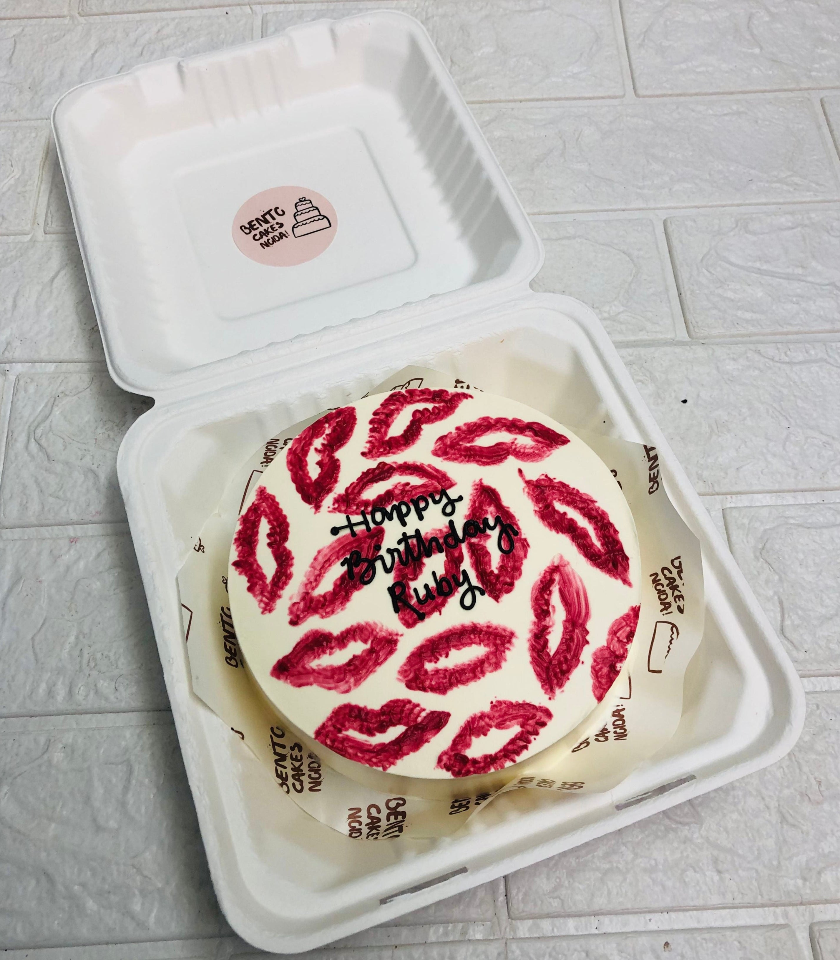 A round birthday cake with white frosting, decorated with multiple red lipstick kiss marks and the message 'Happy Birthday Ruby' written in black icing.