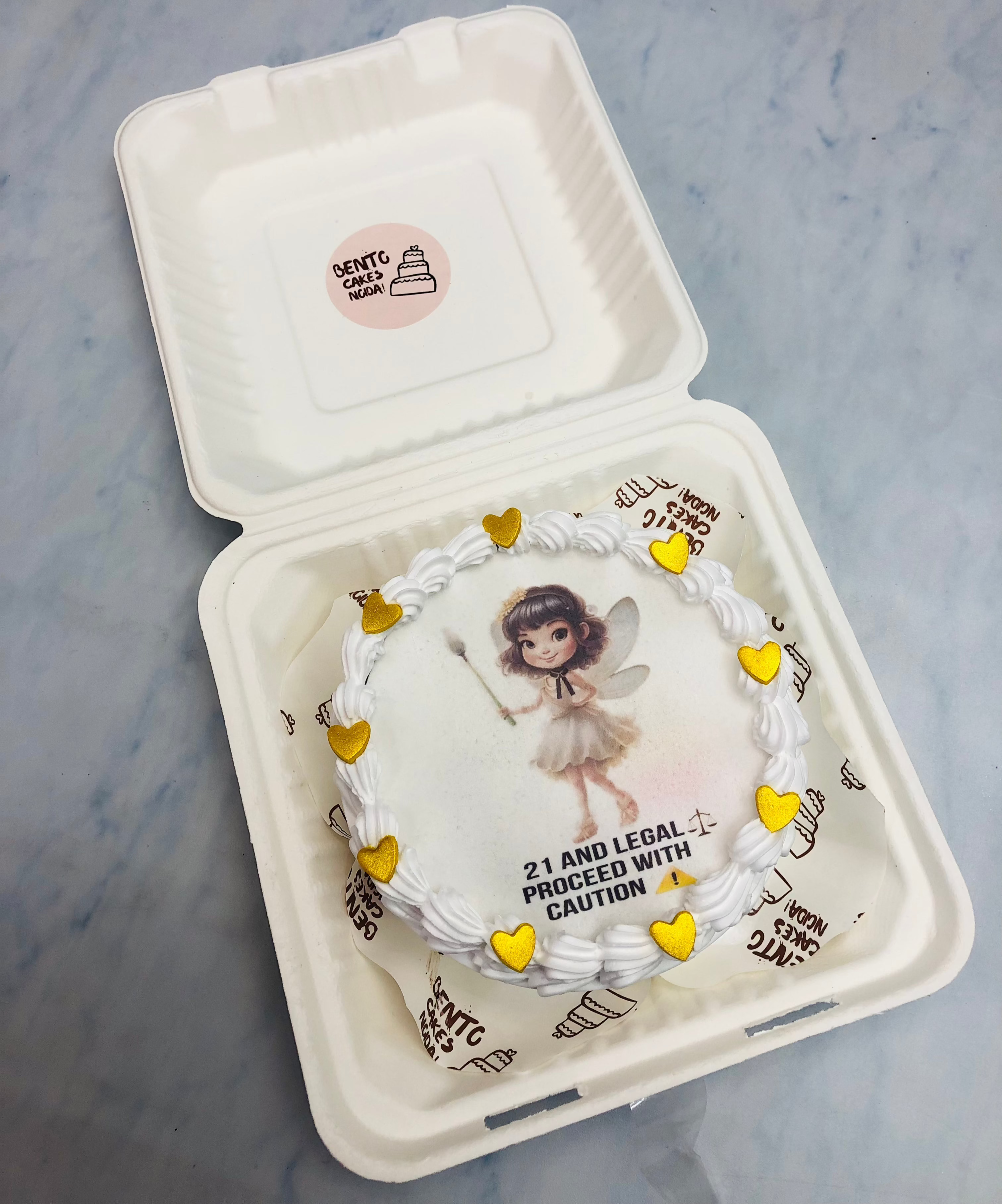 A white fire bento cake with a angel print on fire paper and decorated with golden hearts on it's boundary.