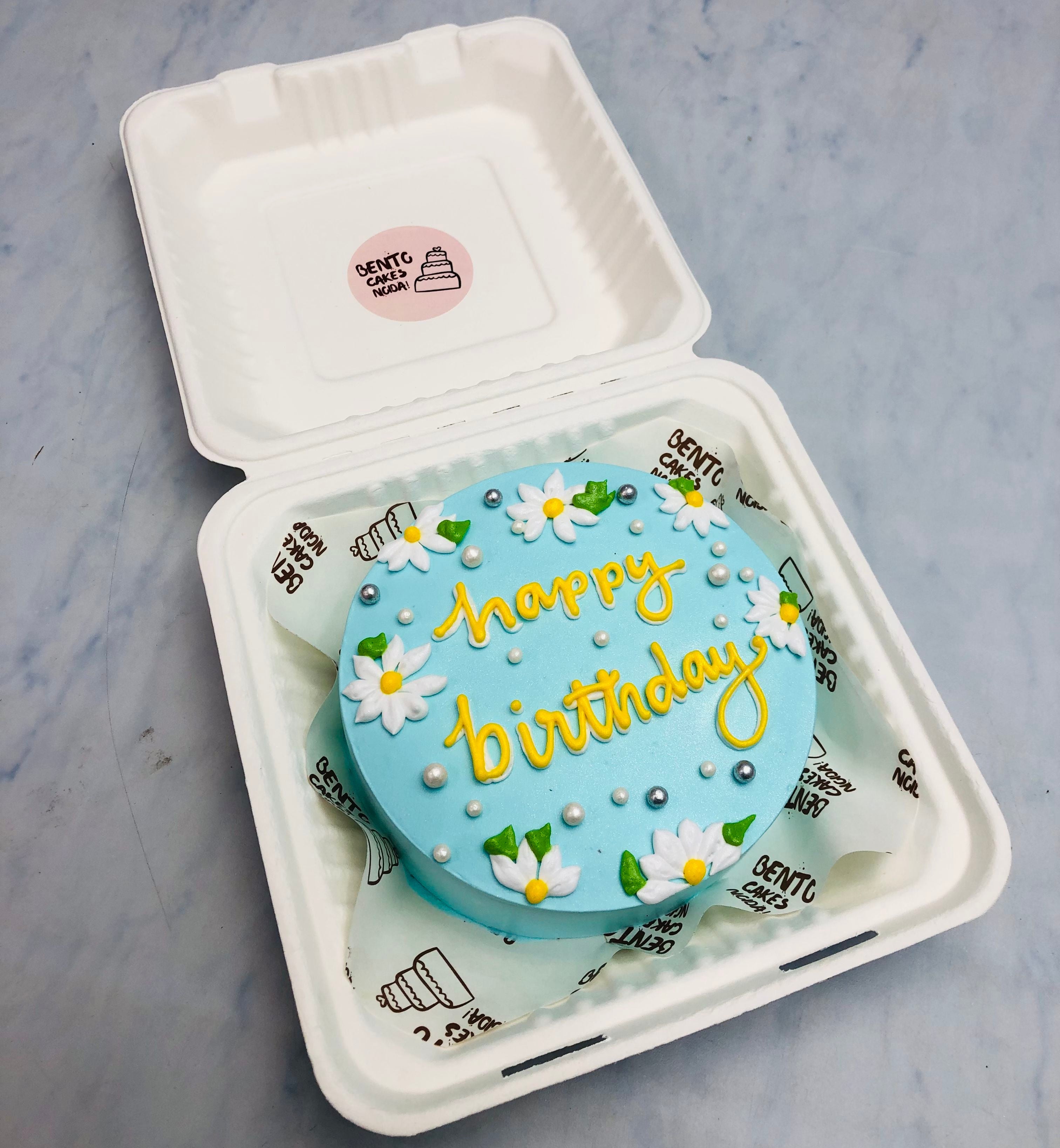 A beautifully crafted blue birthday cake inside a white box, decorated with white daisy flowers, silver pearls, and yellow 'happy birthday' icing.