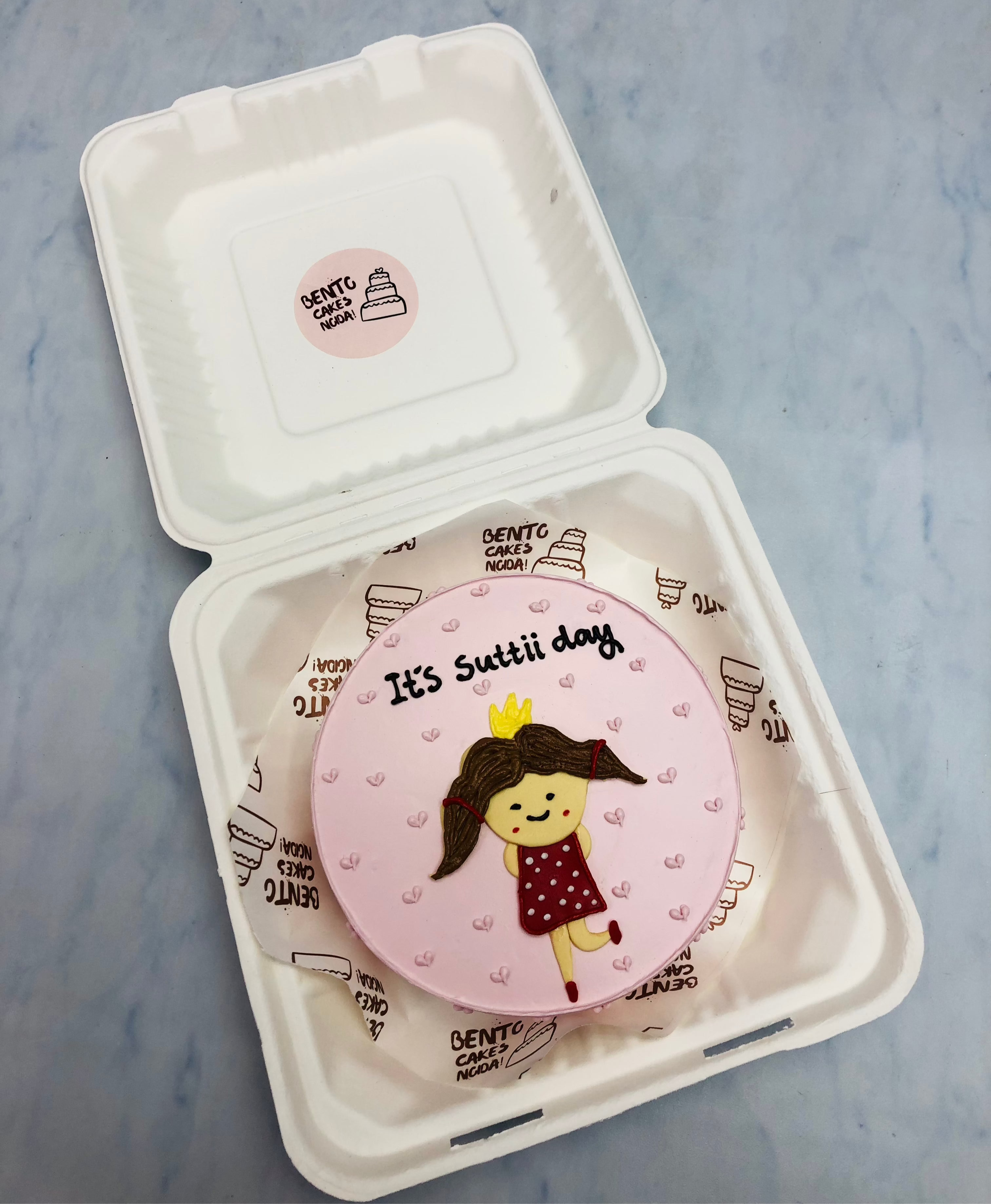 A delightful pink bento cake inside a white container, decorated with an adorable illustration of a girl in a red polka-dot dress and yellow crown. The cake is adorned with small hearts and the playful phrase “It’s Suttii day.”