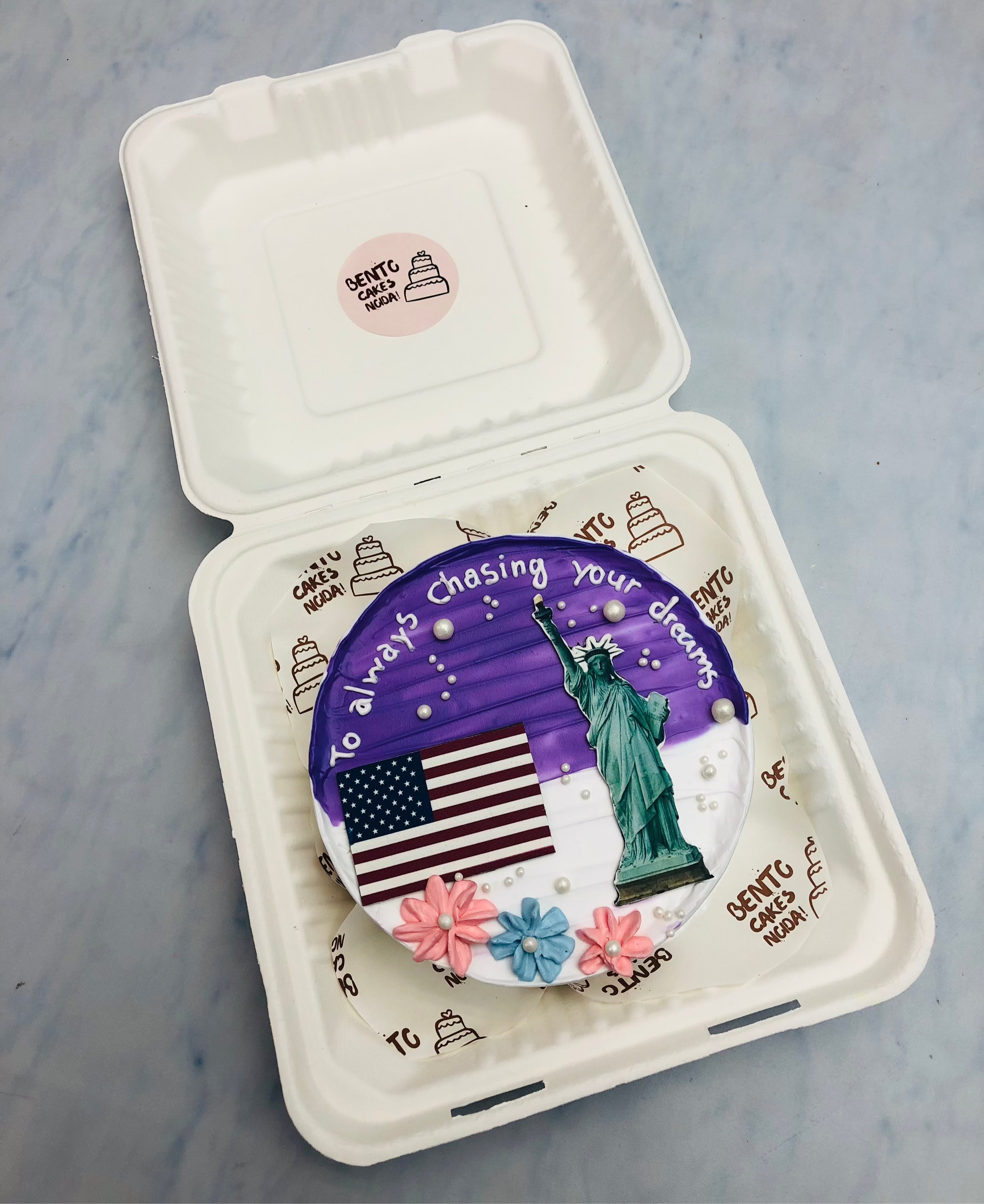An aesthetic blue-white bento cake with a statue, american flag design and decorated with florals.