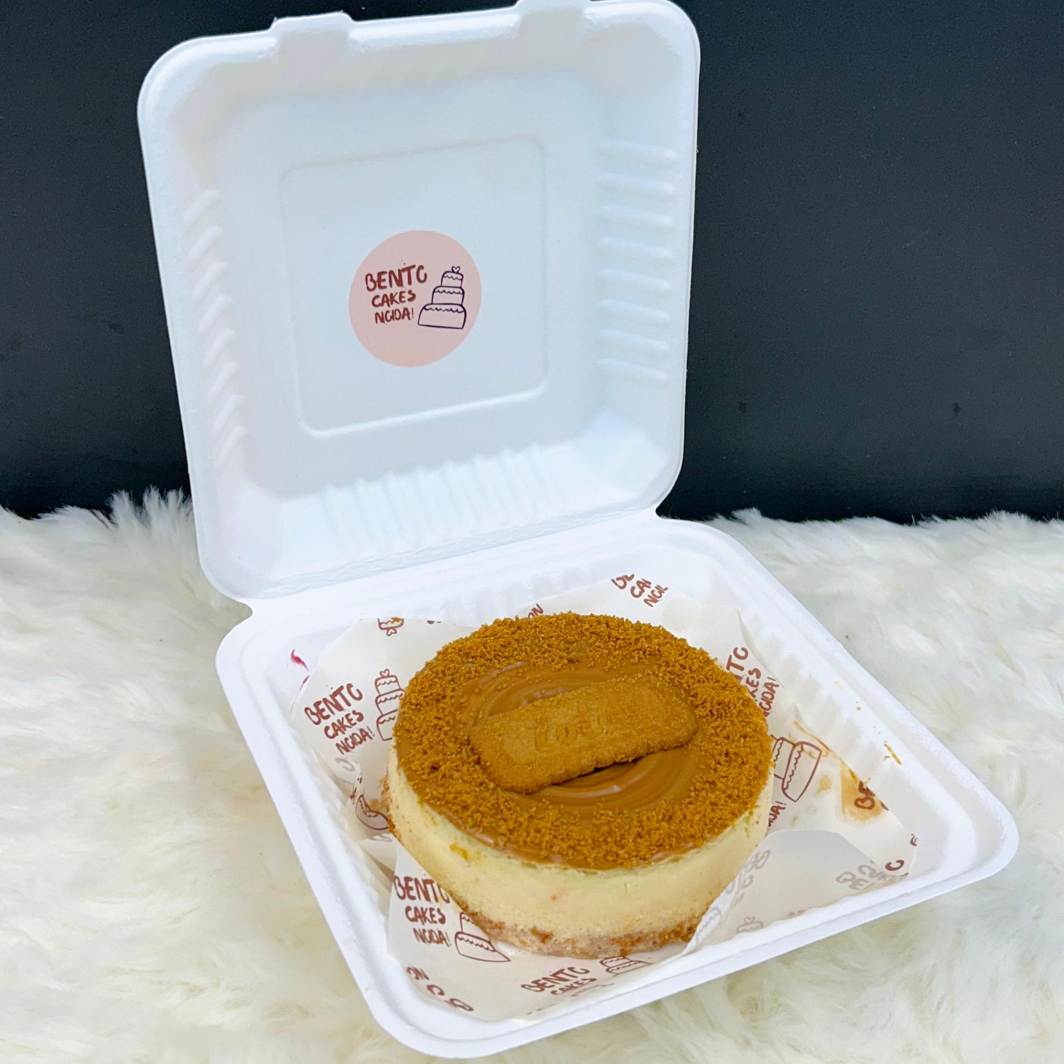 An elegant Lotus Biscoff Cheesecake Bento in a white takeaway container, topped with a smooth layer of Biscoff spread, crushed Biscoff cookies, and edible golden accents on a premium parchment liner.