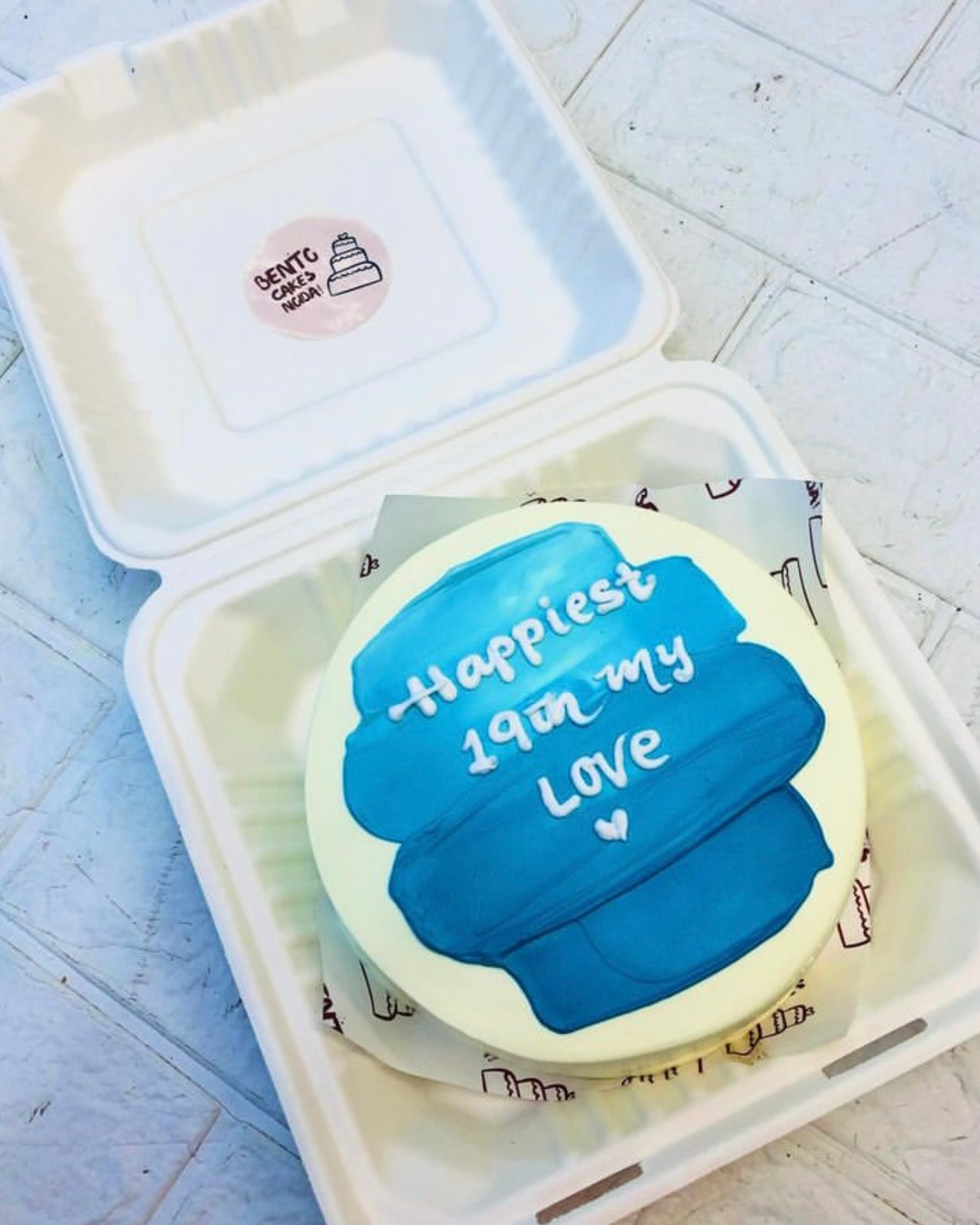 An off white bento cake having a blue shades on top and a text written with whie icing.