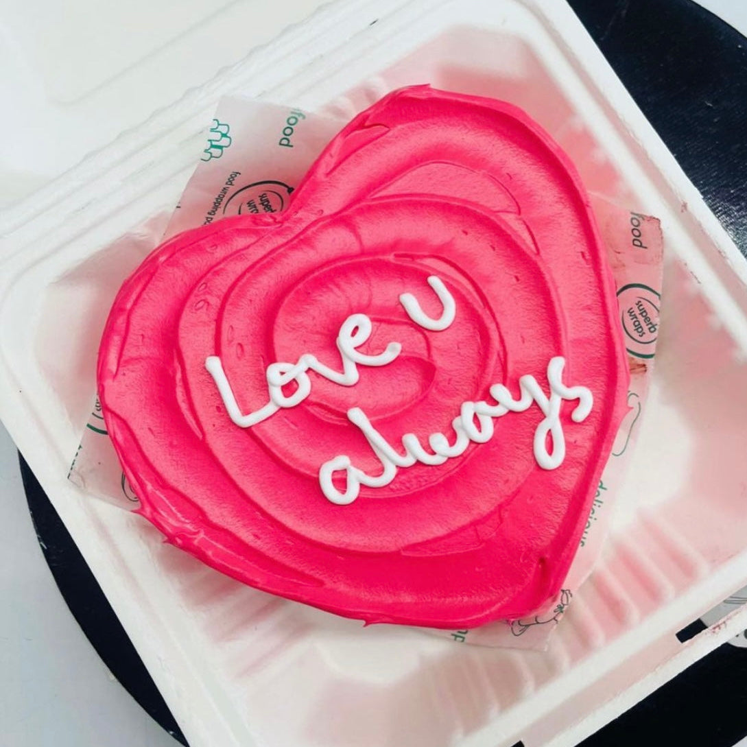 A pink heart-shaped cake adorned with the words "Love U Always" elegantly written on its surface.