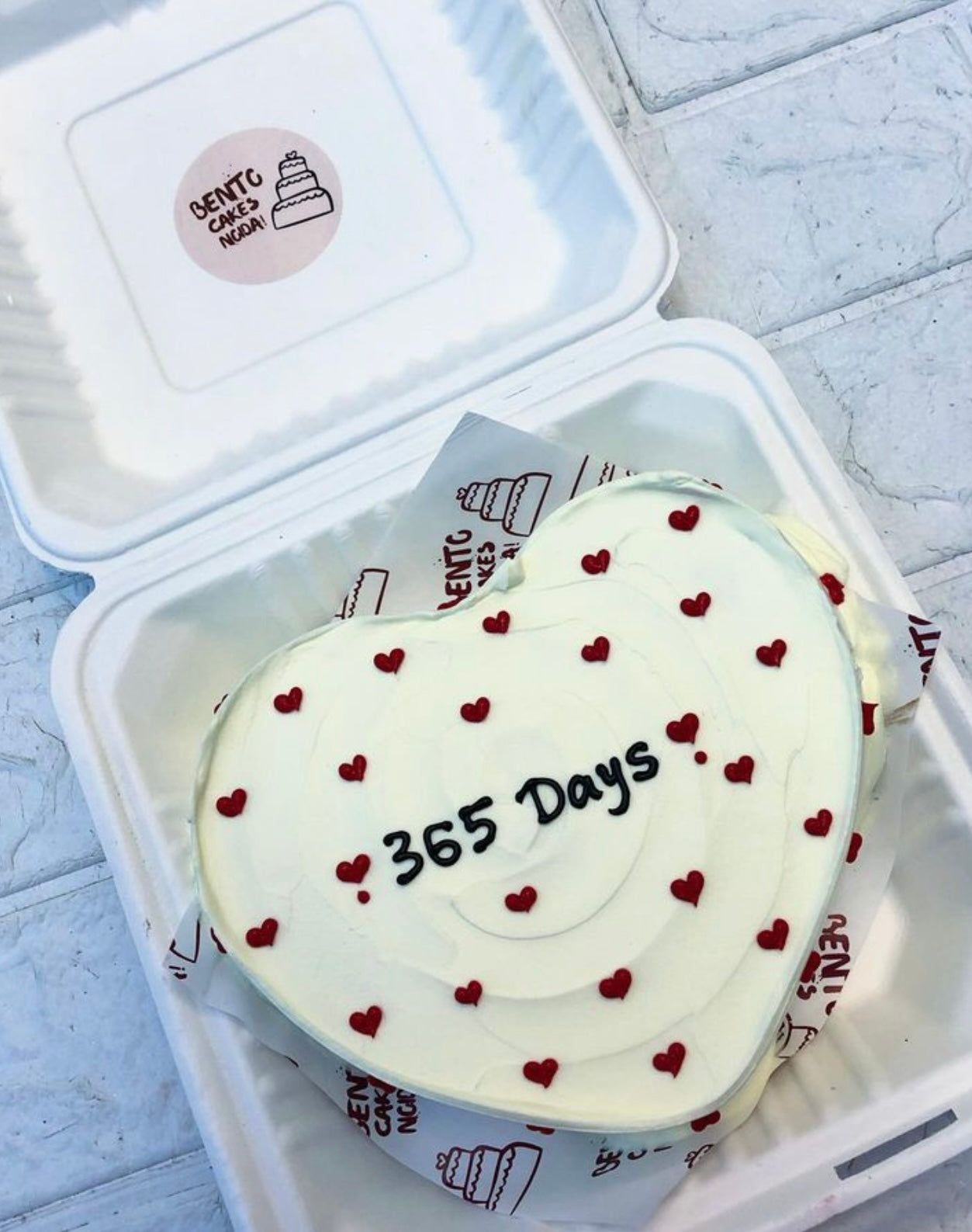 An elegant white cake with red hearts all over the cake and a "365 Days" text written with black icing on it.
