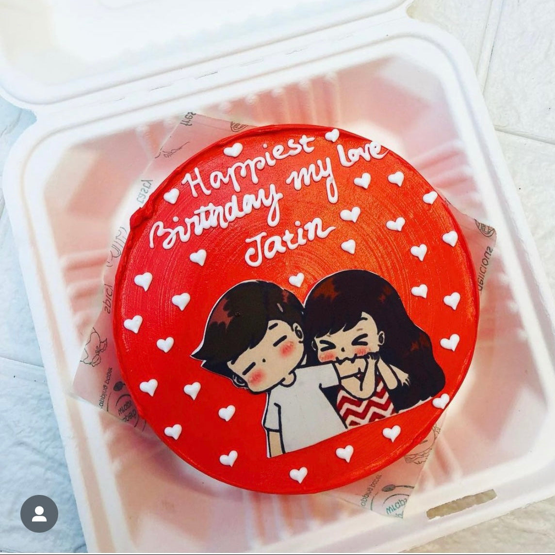 A round red birthday cake with white icing text that reads "Happiest Birthday my love Jatin" and decorated with small white hearts. The cake features a cute illustration of a couple, with the girl playfully pinching the boy's cheek. The cake is placed inside a bento box.